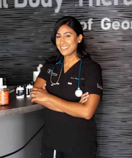 A smiling female healthcare professional wearing scrubs and a stethoscope standing in a clinic or medical office.