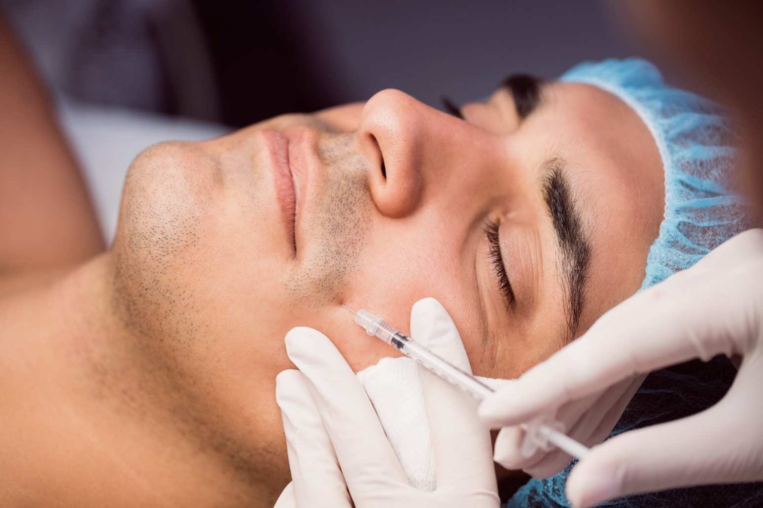 A man receiving a cosmetic injection on his face while lying down, wearing a blue surgical cap, with healthcare professional's gloved hand holding a syringe.