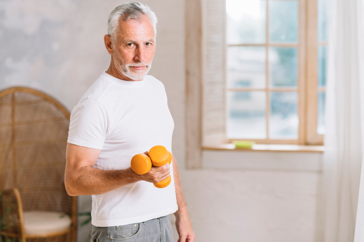 Older man with gray hair and beard holding yellow dumbbell, standing in a bright room with a window in the background.