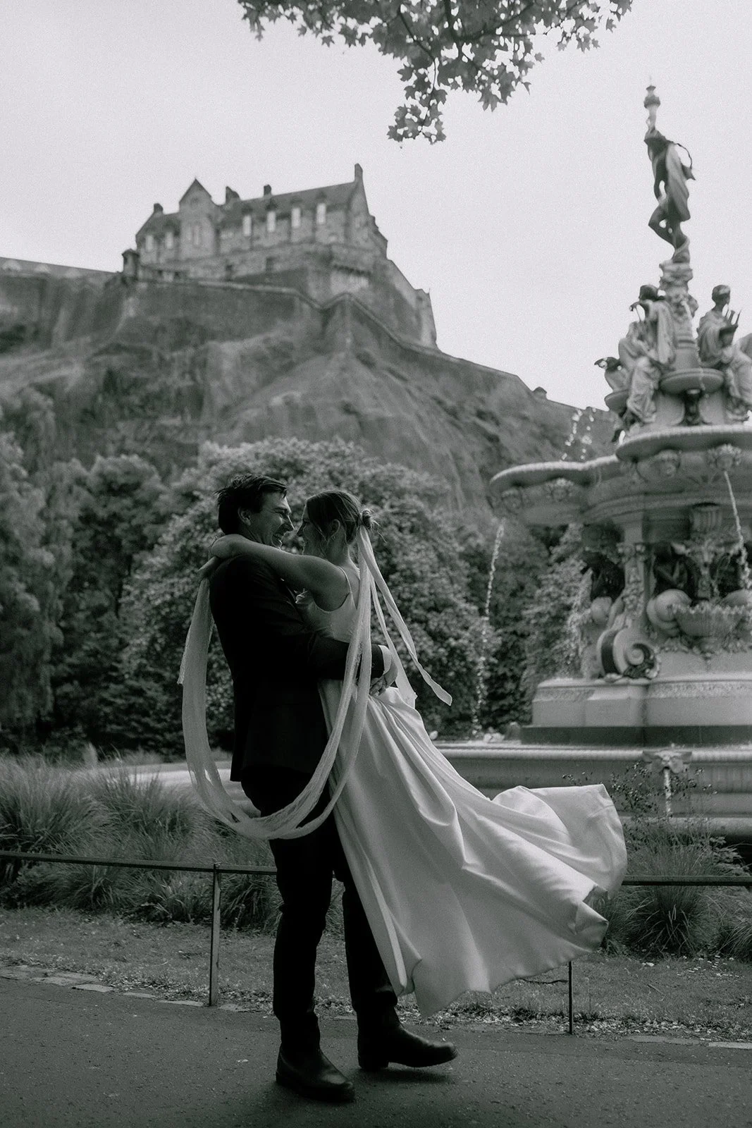 A wedding portrait of bride and groom dancing in front of a fountain and a castle on a hill, with one person lifting the other in a romantic pose in Edinburgh Princes street gardens for their wedding elopement