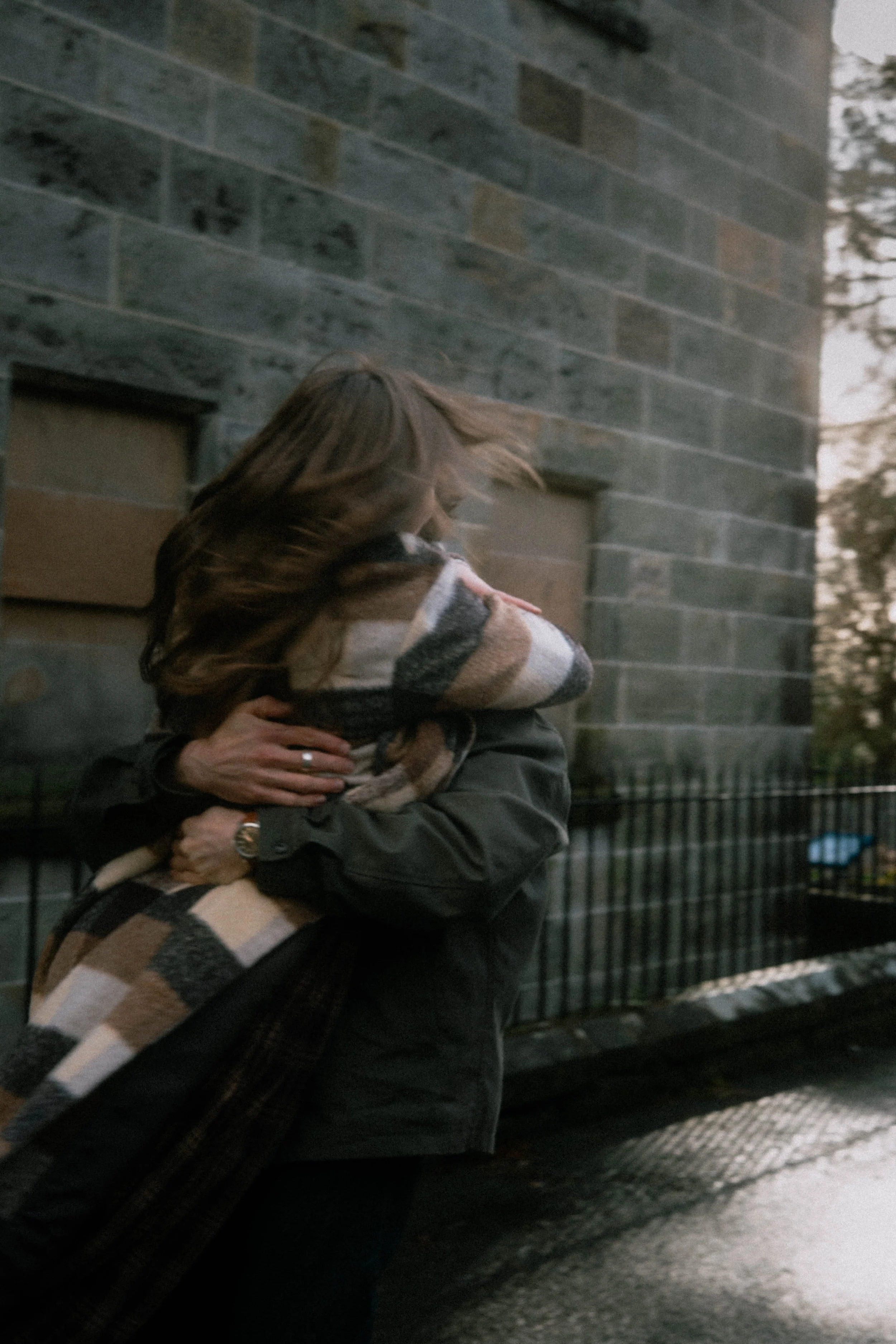 Person with brown hair holding a cat wrapped in a plaid blanket, standing outdoors near a brick building, with trees in the background during evening or overcast weather.