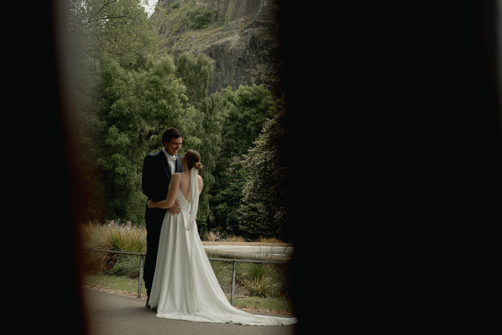 A bride and groom are standing close together outdoors, the groom in a tuxedo and the bride in a white wedding dress, looking lovingly at each other, framed by a small opening in princes street. natural Edinburgh elopement and wedding photography.