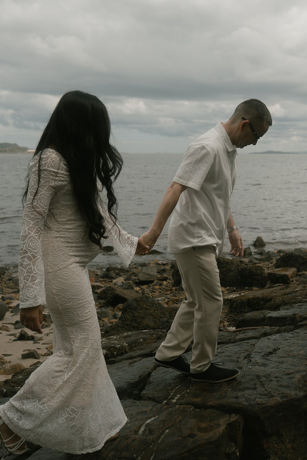 A man and woman holding hands walking on rocks near the water on a cloudy day, the woman wearing a white lace dress and the man wearing light-colored clothing and sunglasses.