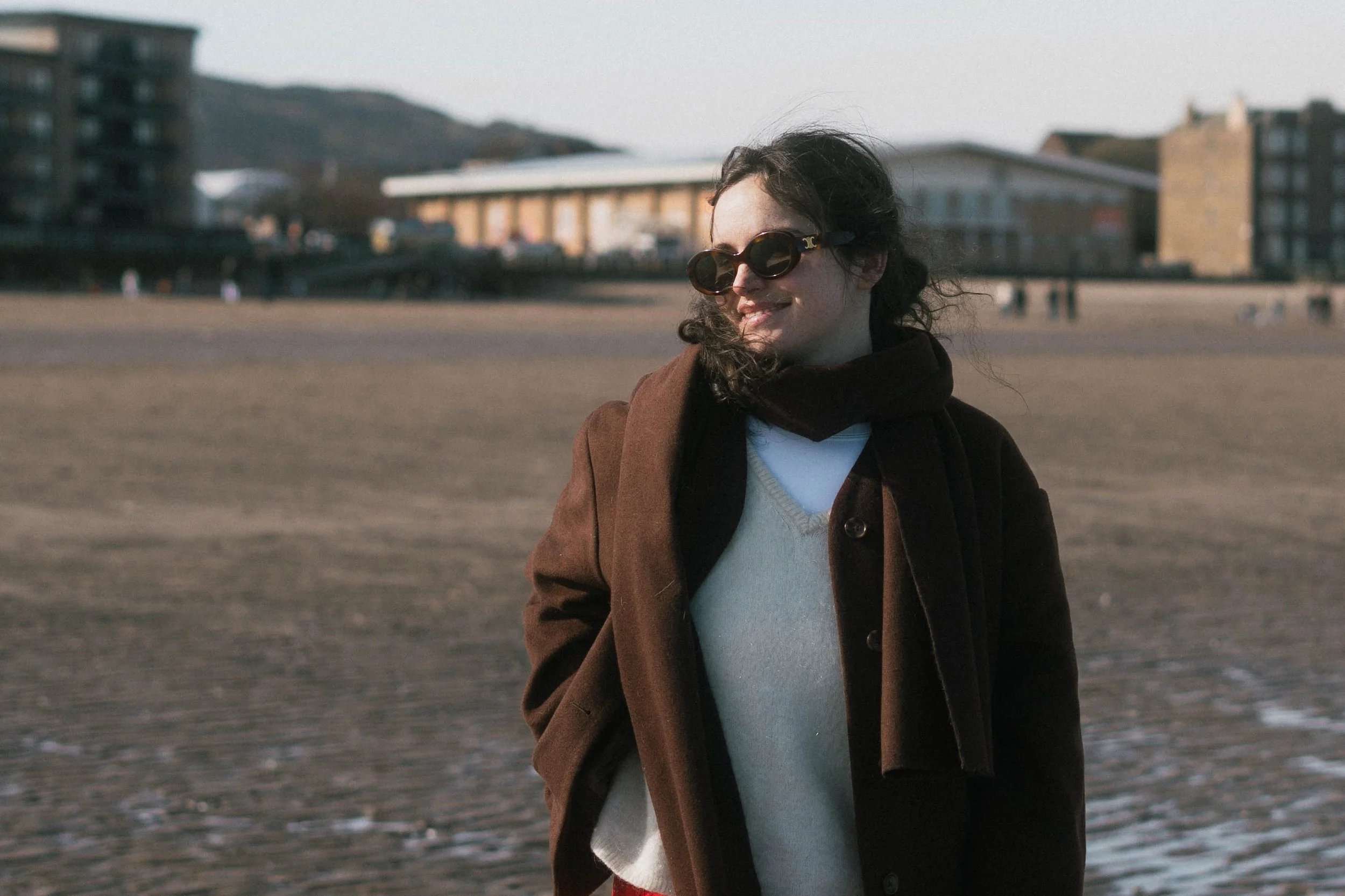 A woman with dark, curly hair smiling and wearing sunglasses, a brown coat, black scarf, and light-colored sweater standing on a beach with buildings and hills in the background.