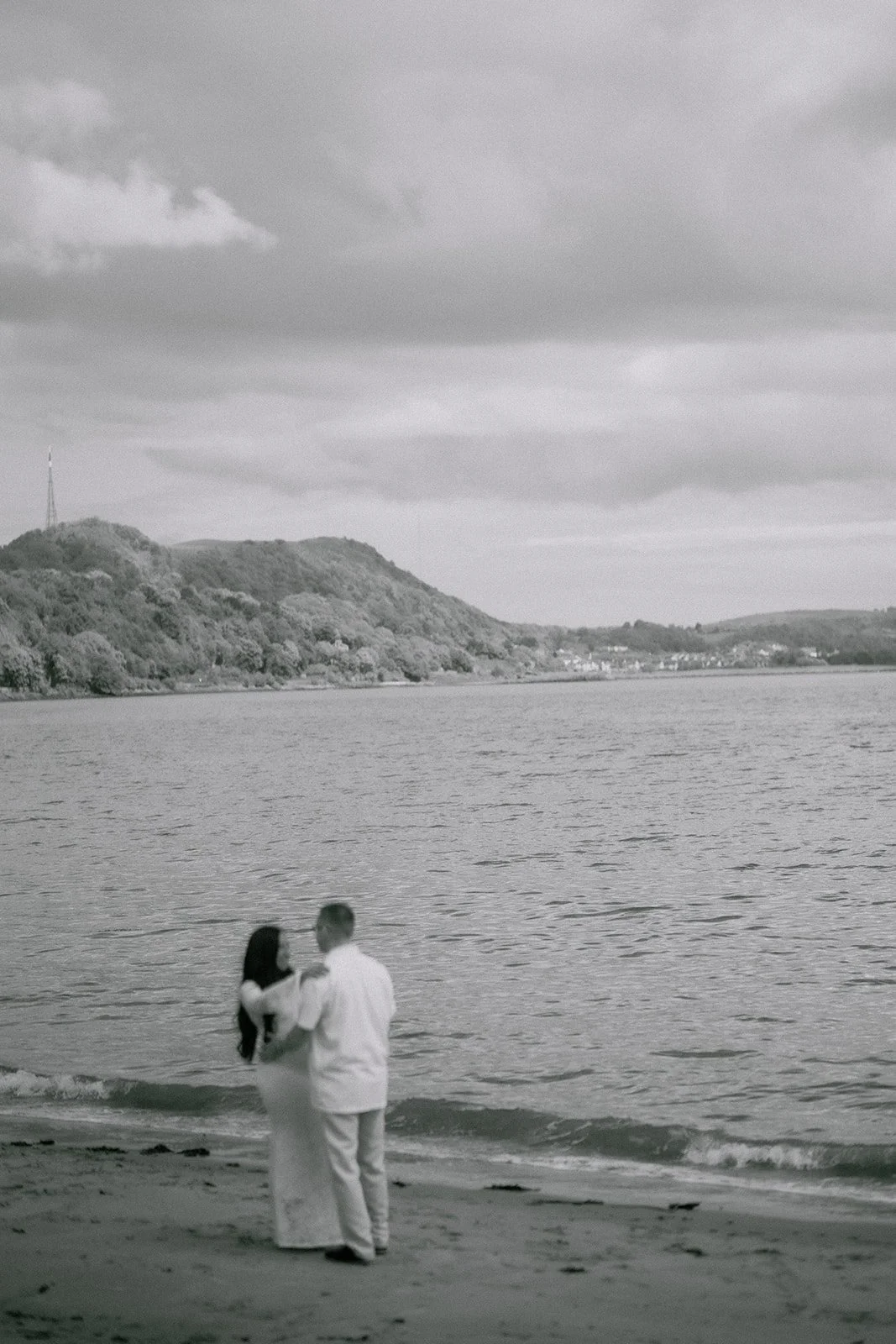 A Pinterest couple standing on a beach holding hands near the water sh in a black and white photograph, with hills and cloudy sky in the background. Captured by Scotland based elopement photographer