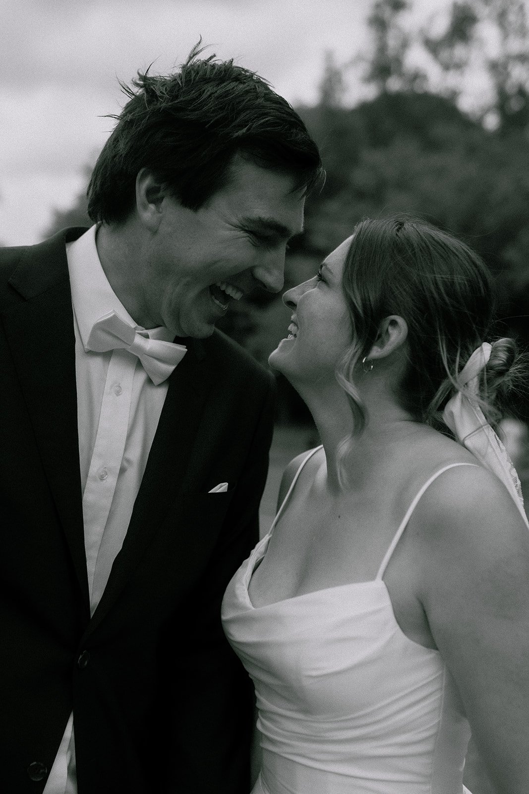 A black-and-white photo of a happy couple smiling at each other outdoors. The man is dressed in a tuxedo with a bow tie, and the woman is wearing a dress with spaghetti straps. Arthurs seat natural Edinburgh elopement and wedding photography.