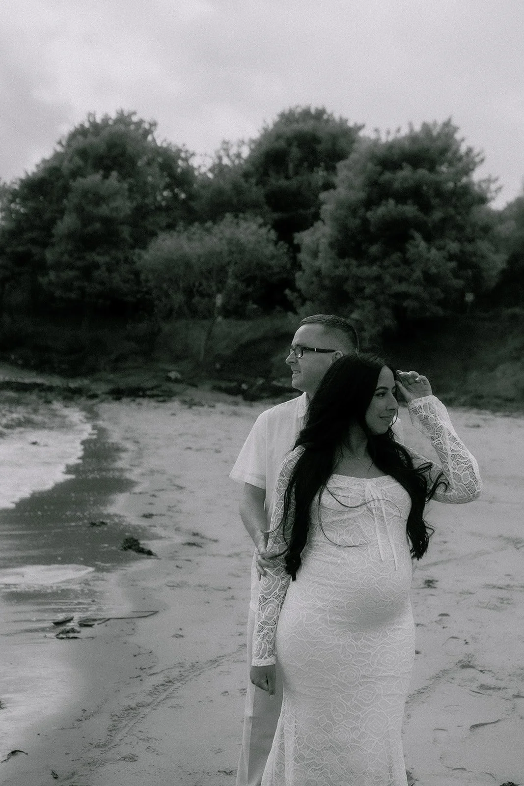 A black-and-white photo of a pregnant woman in a lace dress standing on a beach, with a man in glasses behind her. They are near water and trees are in the background. A natural and candid maternity and couples photoshoot in Fife, Central Scotland.