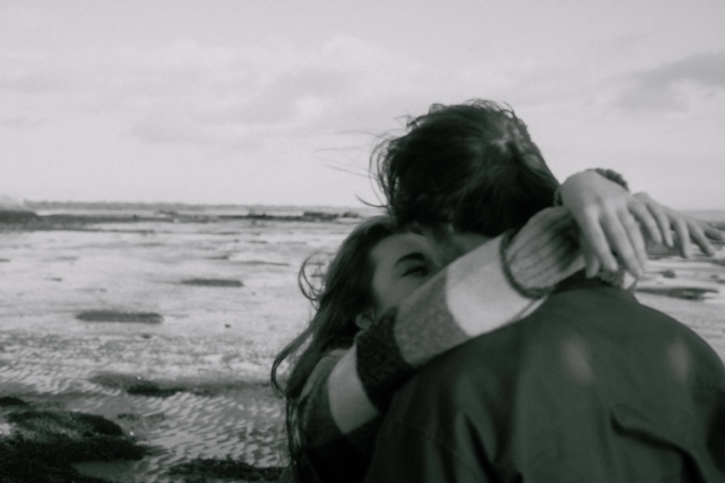 Two people hugging on a beach in Culross, Fife in Scotland, one with long hair and the other person's face not visible, in black and white, slightly blurred with a Pinterest couples vibe.