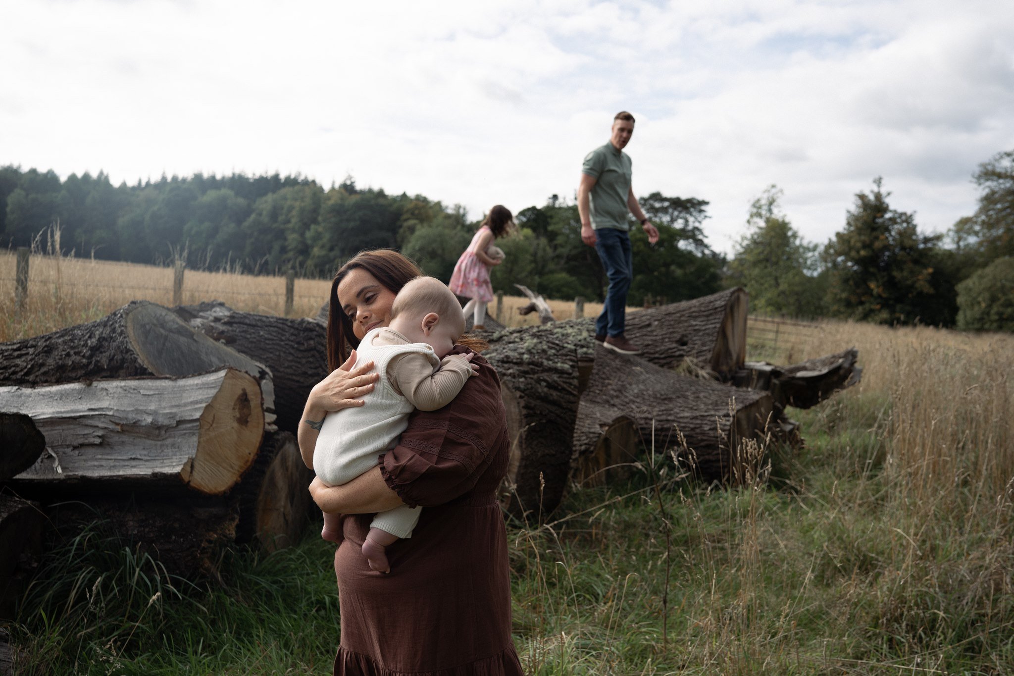 a natural and candid photo of a mum with baby and dad with little girl climbing on logs in Falkland estate in Scotland fife for their family photoshoot