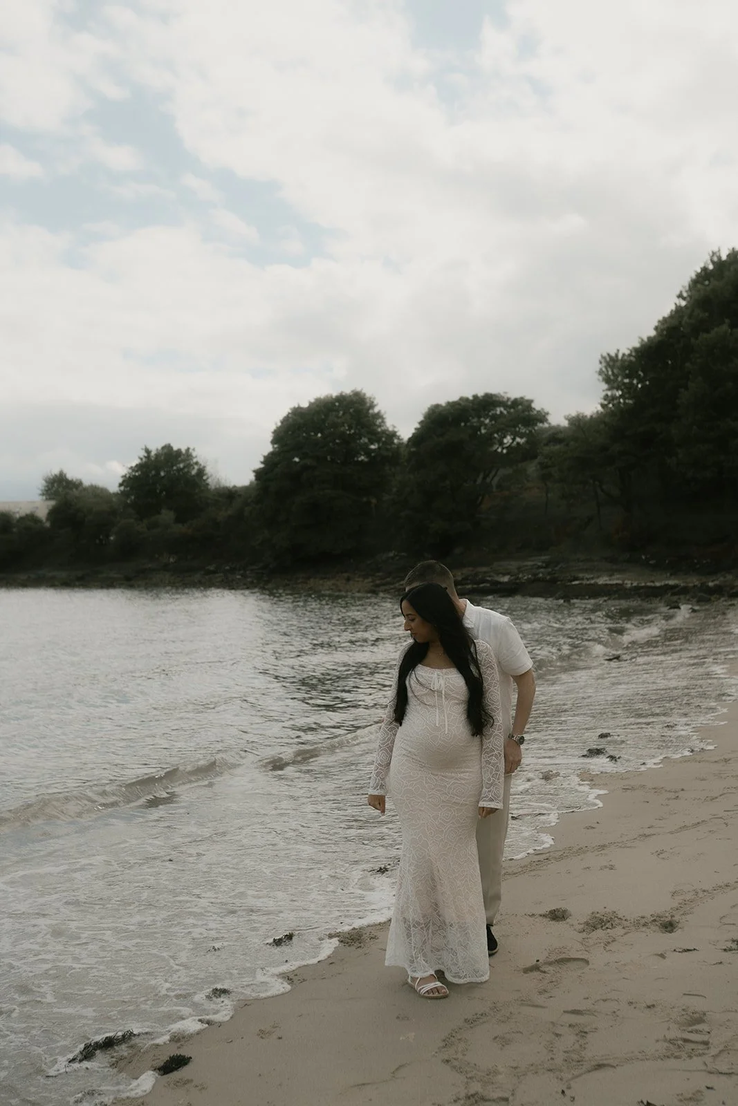 A couple during their natural maternity photoshoot on a in Aberdour, Fife in Scotland. They're walking on a beach, with the woman in a white dress and the man behind her, looks like Scottish elopement photography.