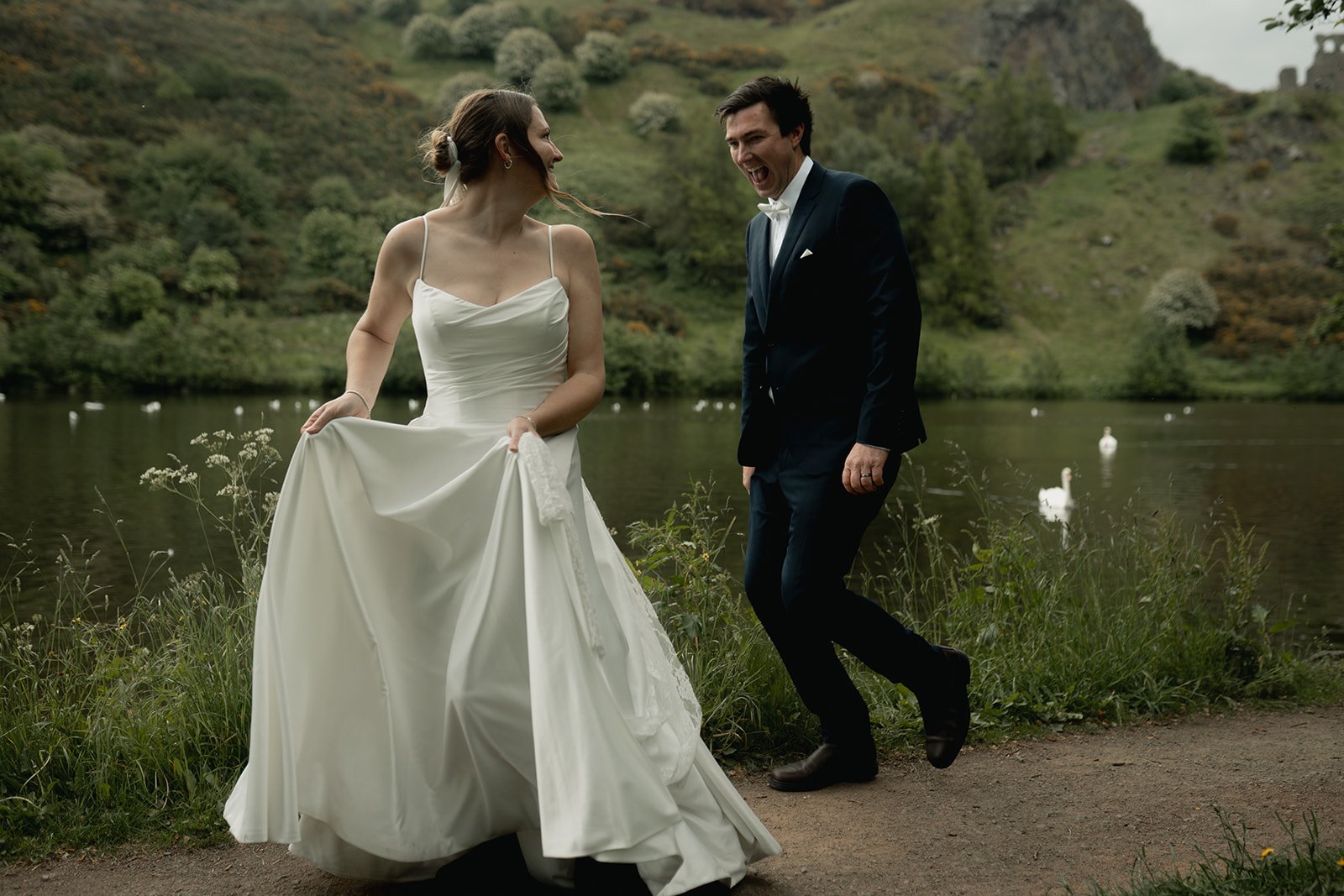 A bride in a white wedding dress and a groom in a dark suit with a bow tie celebrate outdoors near a lake with swans, surrounded by green hills and trees. Hollyrood park elopement, natural and candid wedding photography.