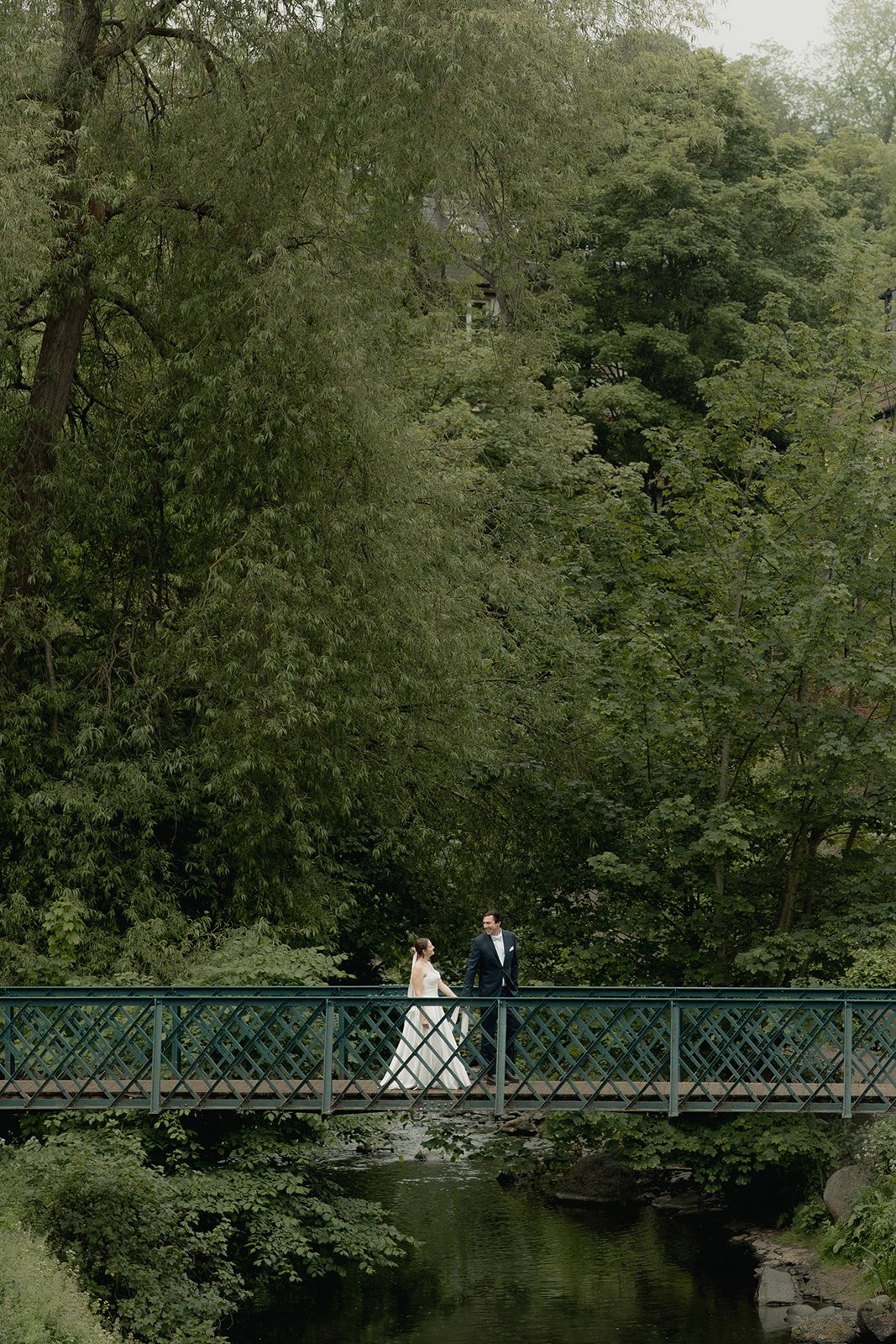 A bride and groom holding hands while walking on a small arched bridge over a creek, surrounded by lush green trees in Edinburgh dean Village for their elopement wedding