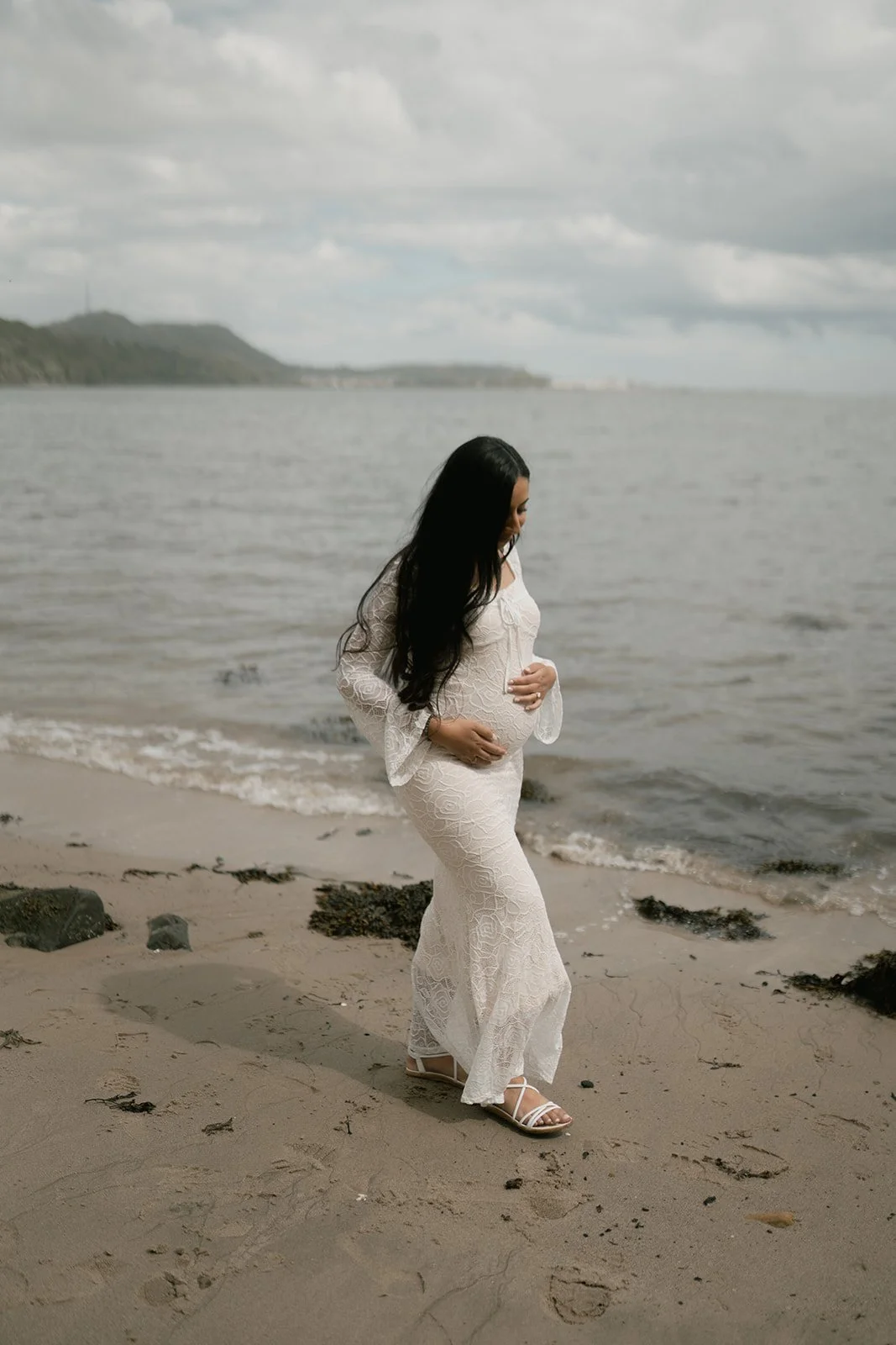 A pregnant woman in a white lace dress standing on a beach, looking down at her belly with her hands on her stomach.