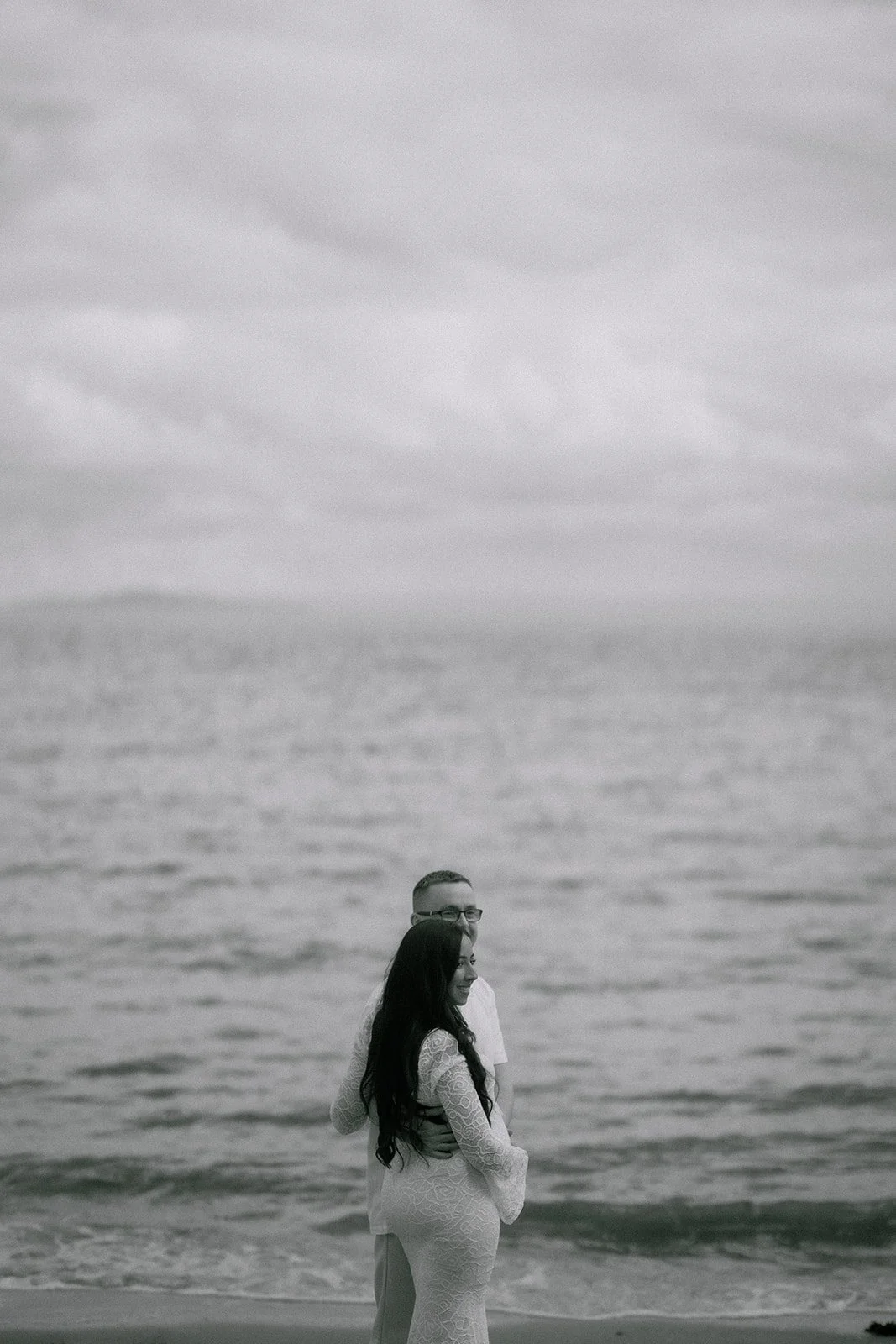 A couple embraces on a beach with ocean waves and cloudy sky in the background. A natural and candid maternity and couples photoshoot in Fife, Central Scotland.