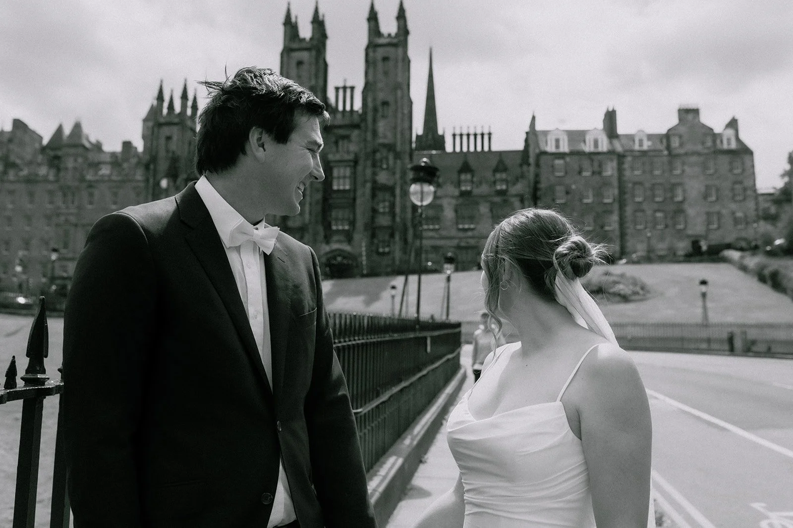 A black and white wedding photo of a man in a tuxedo and a woman in a white dress standing outdoors near a castle or historic building in Edinburgh, smiling and looking at each other during their elopement photoshoot.