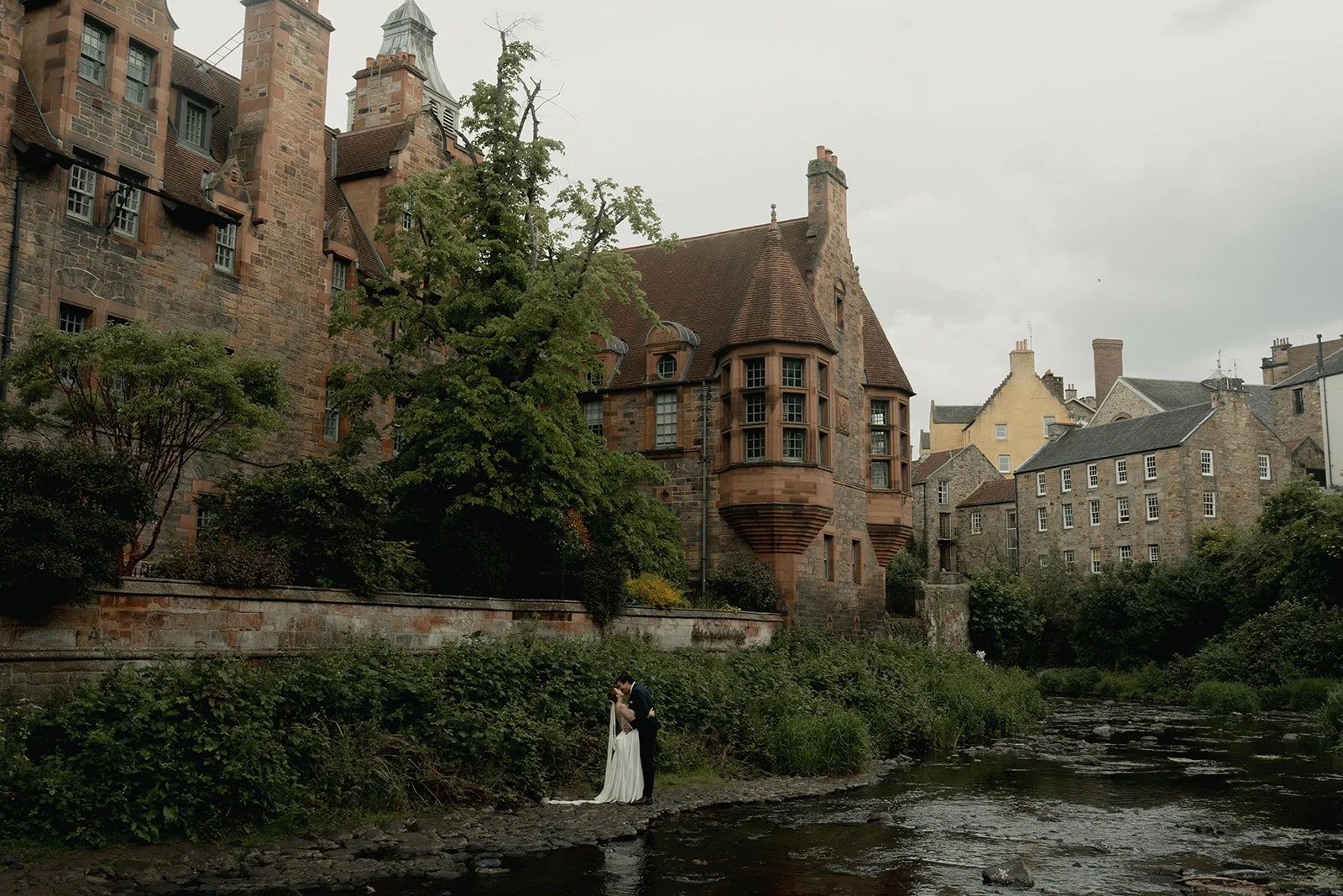 A groom and bride are standing on a riverbank, kissing, with a historic stone building with turrets and multiple windows in the background, surrounded by greenery and overcast sky.