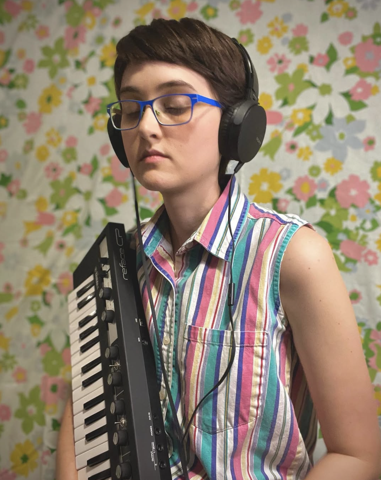 Person with short brown hair, wearing blue glasses and headphones, holding a compact keyboard, in front of a colorful floral background.