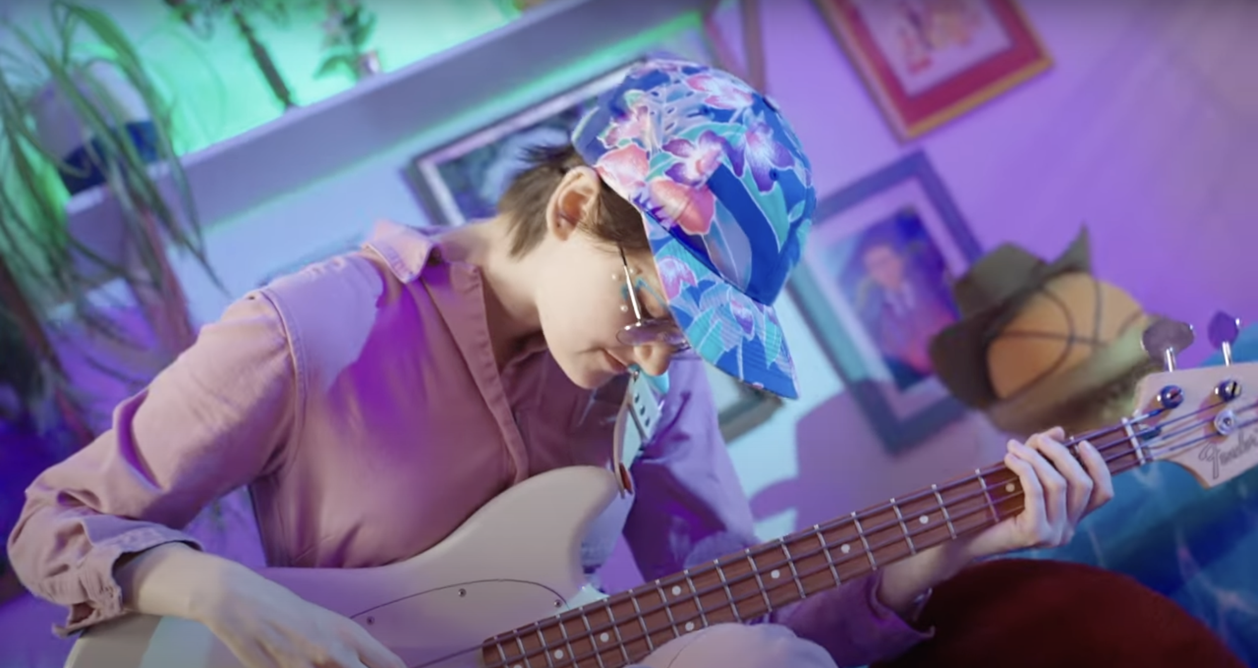 A person wearing a colorful cap, pink shirt, and glasses playing an electric bass guitar in a room decorated with framed pictures, plants, and colorful lighting.