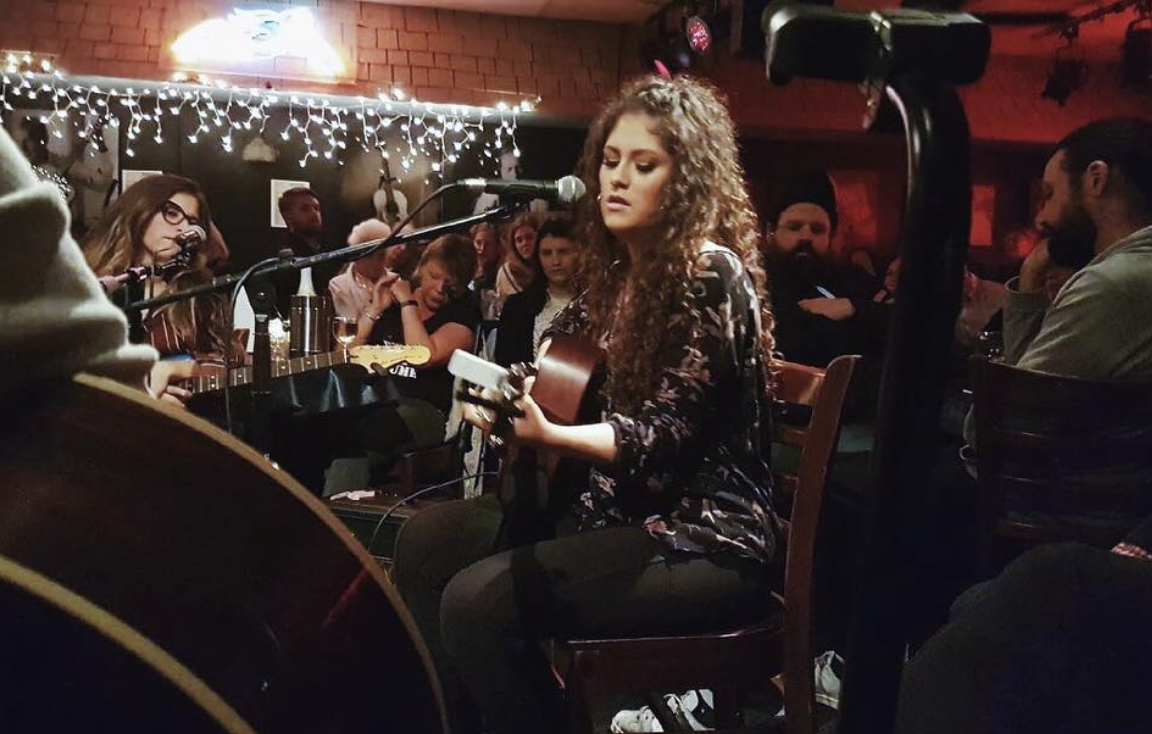 A woman with curly hair playing an acoustic guitar during a live music performance at a cozy venue, with audience members watching attentively and fairy lights decorated in the background.