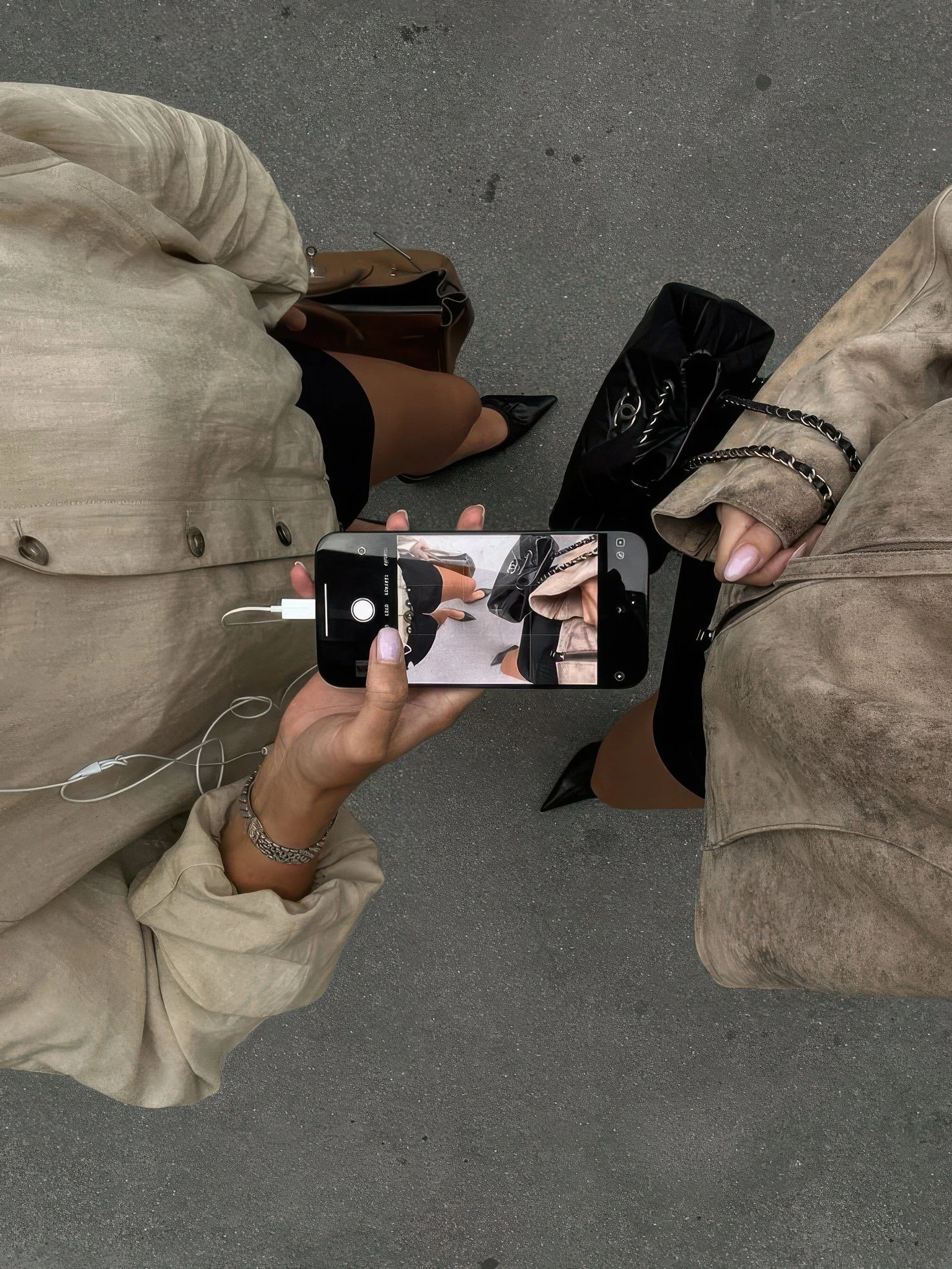From above view of two people standing on a gray asphalt pavement, one person taking a photo with a smartphone, capturing their own camera and part of their beige jacket, black skirt, and shoes. The other person is wearing a beige trench coat, black bag, and pointy shoes, holding a black bag on the ground.