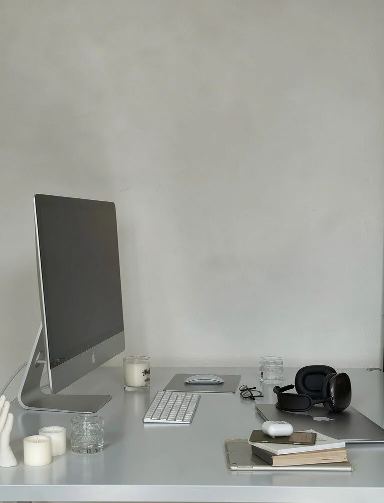 A minimalist white desk with a large iMac computer, a candle, a glass of water, a wireless keyboard, mouse, eyeglasses, a headset, a laptop, a notebook, a smartphone, a pair of wireless earbuds, and some candles.