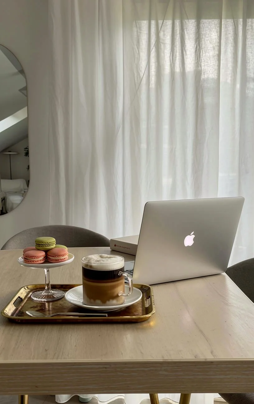 A wooden table with a tray holding a glass of layered coffee, a plate with a spoon, and a stand with three macarons. A closed MacBook laptop and a box are also on the table. Light curtains are in the background.