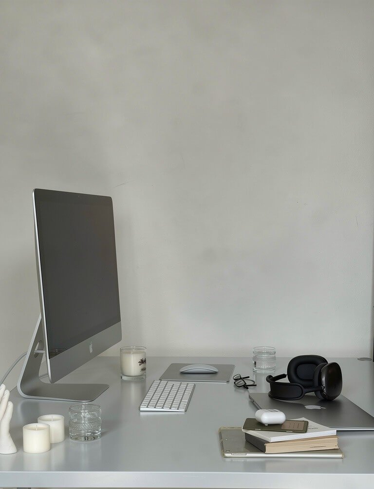 Organized desk with iMac computer, candles, glasses, headphones, glasses, earbuds, notebooks, and a closed laptop.