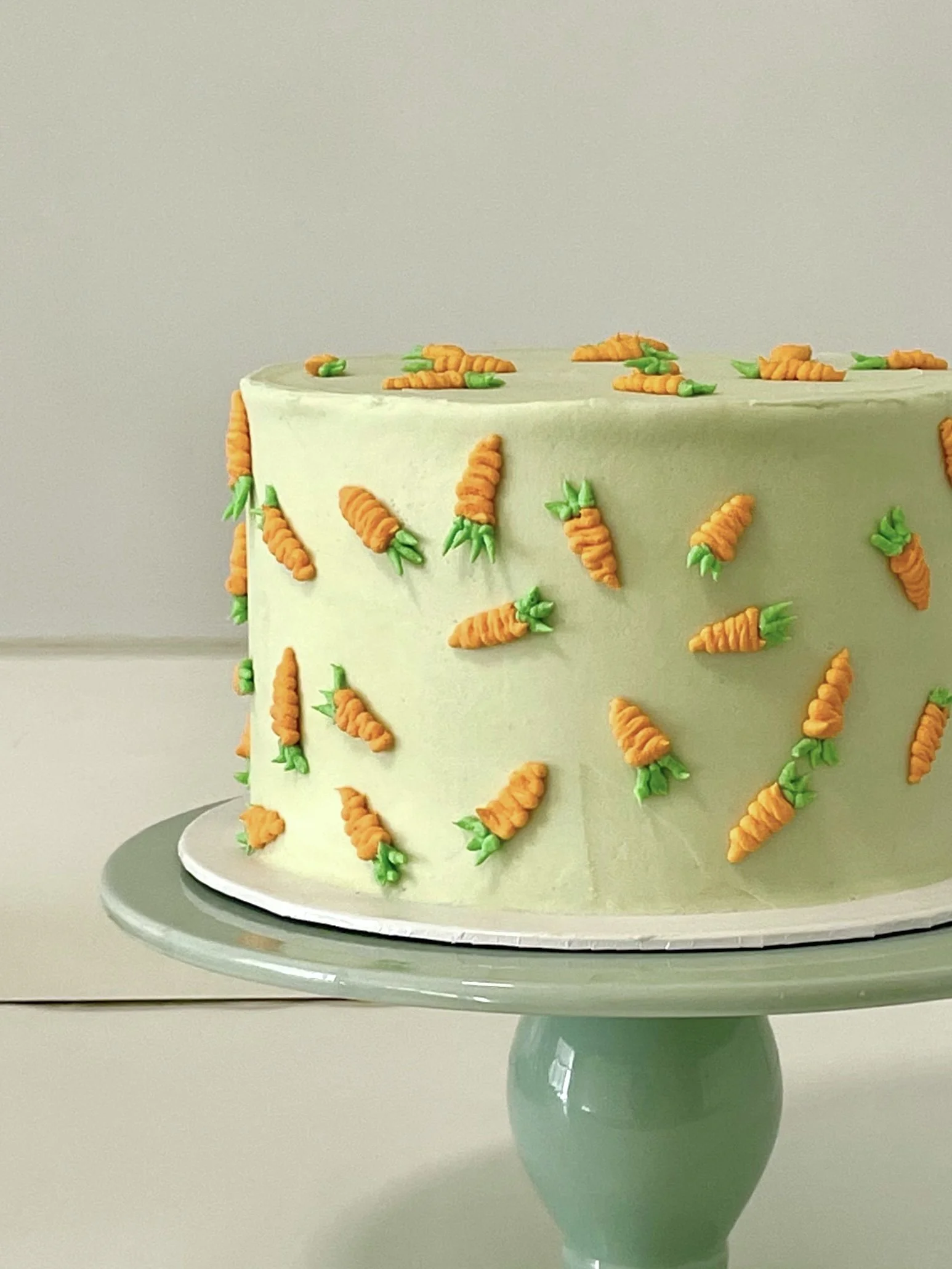 A round cake decorated with small, orange carrot-shaped frosting decorations with green leaves, set on a light-colored cake stand.