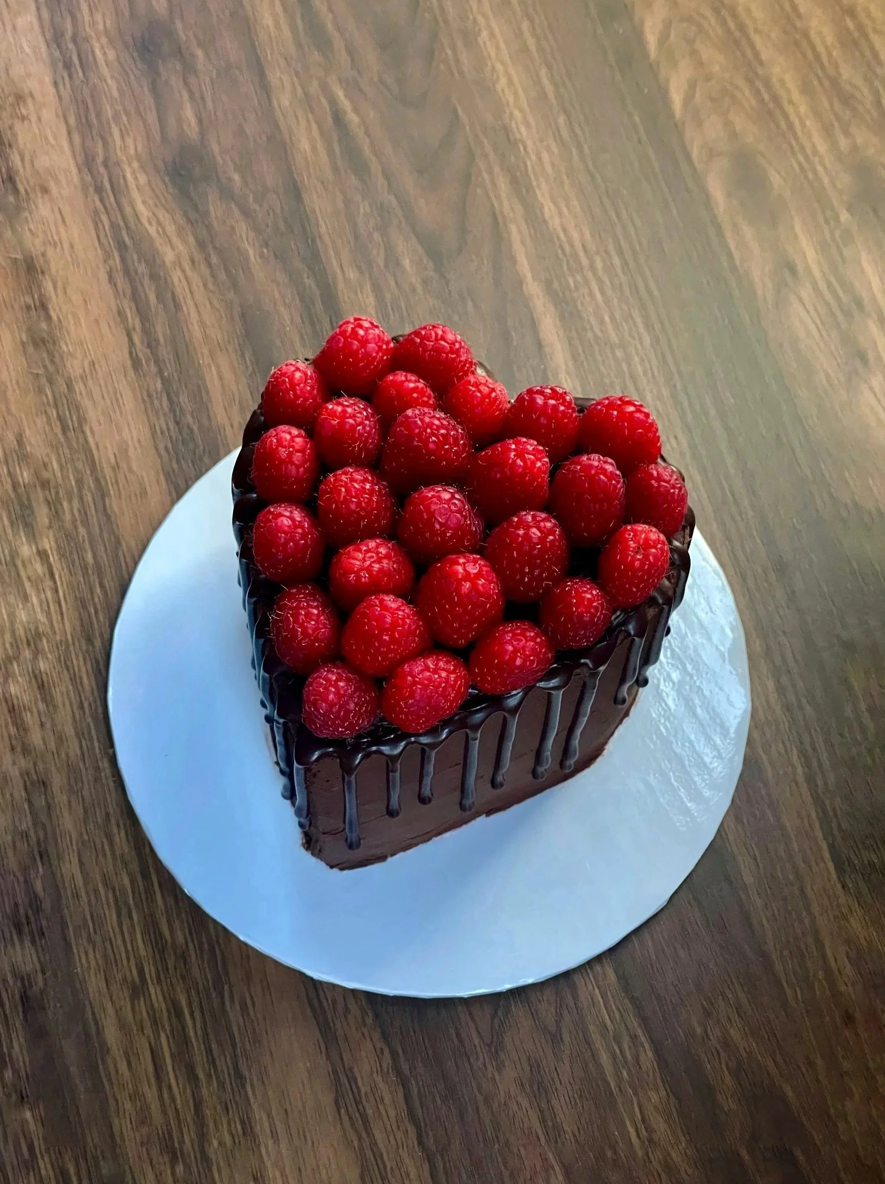 A heart-shaped chocolate cake topped with fresh red raspberries, with chocolate drizzle along the sides, placed on a white round cake board on a wooden surface.