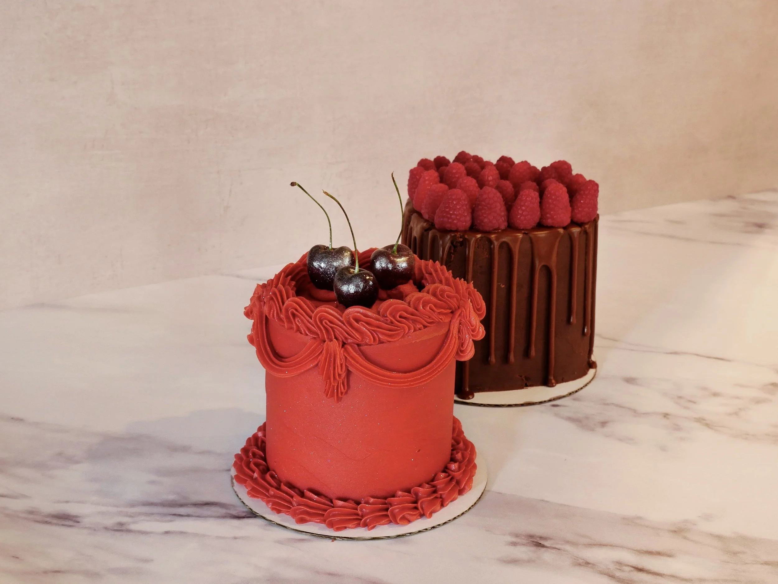 Two decorated cakes topped with fresh berries, cherries, and chocolate drizzle, displayed on a marble surface.