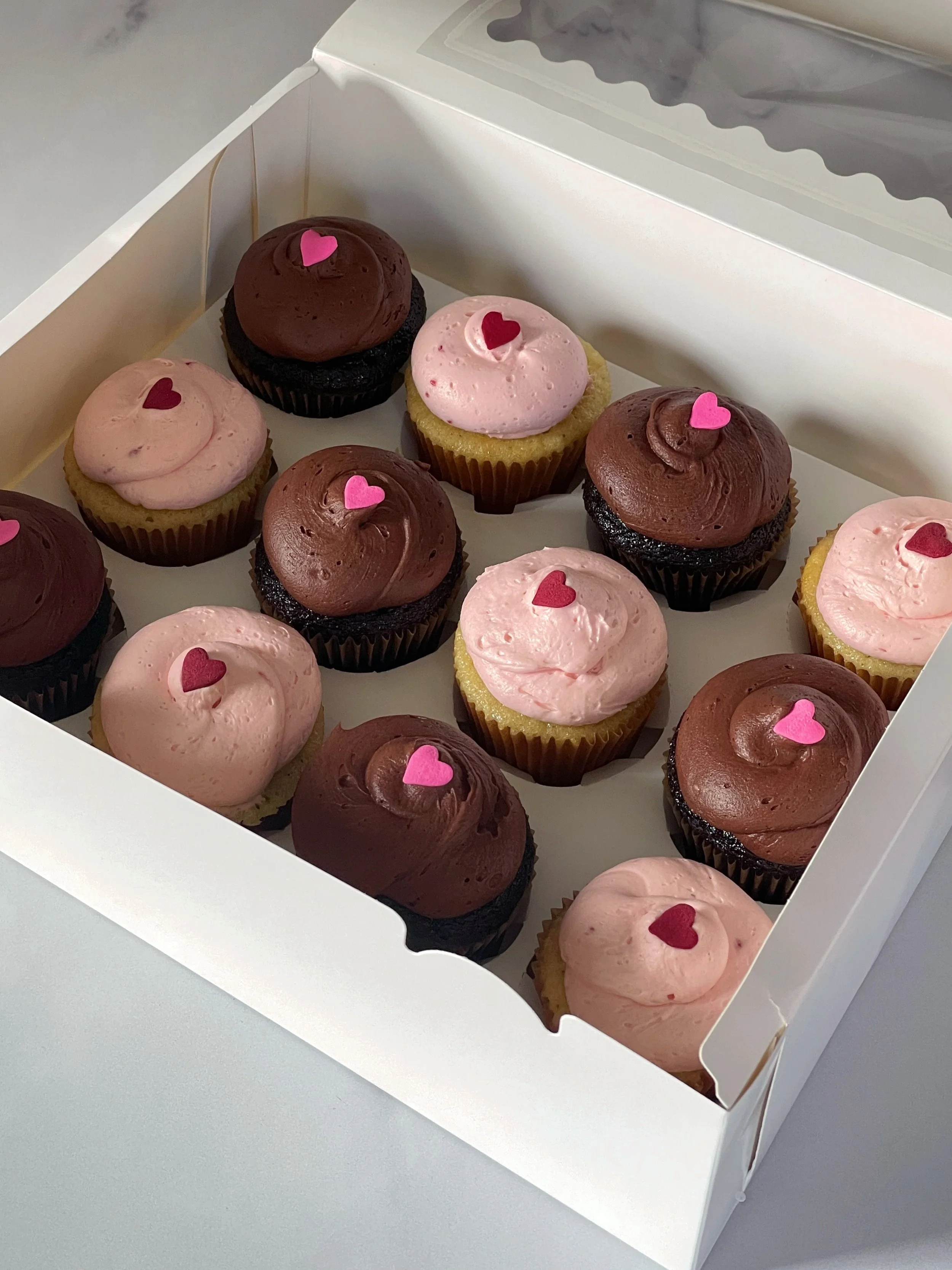 Box of assorted chocolate and vanilla cupcakes with pink and red heart-shaped decorations.