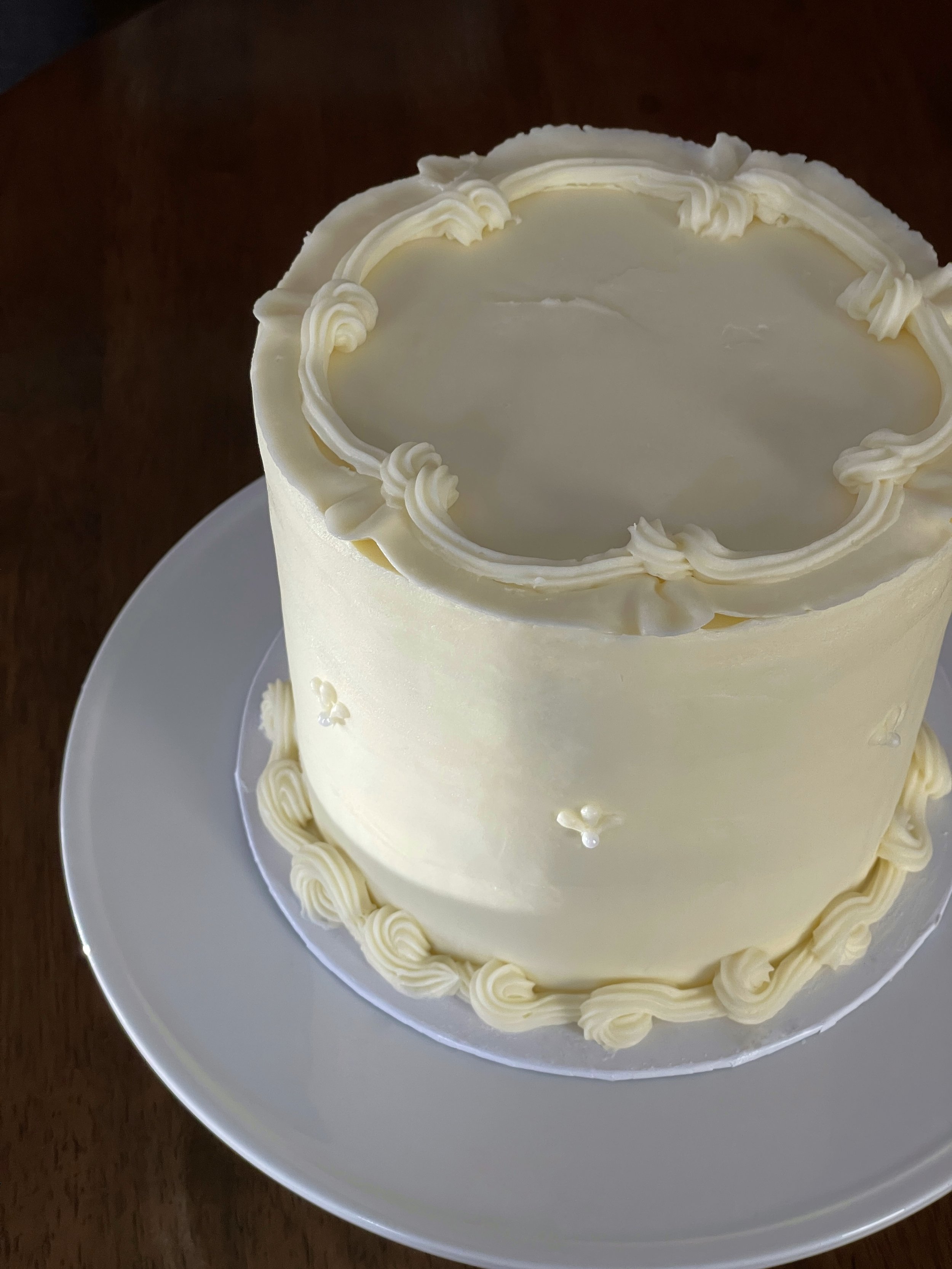 Cream-colored, tall birthday cake with decorative icing on top and around the base, placed on a white plate.