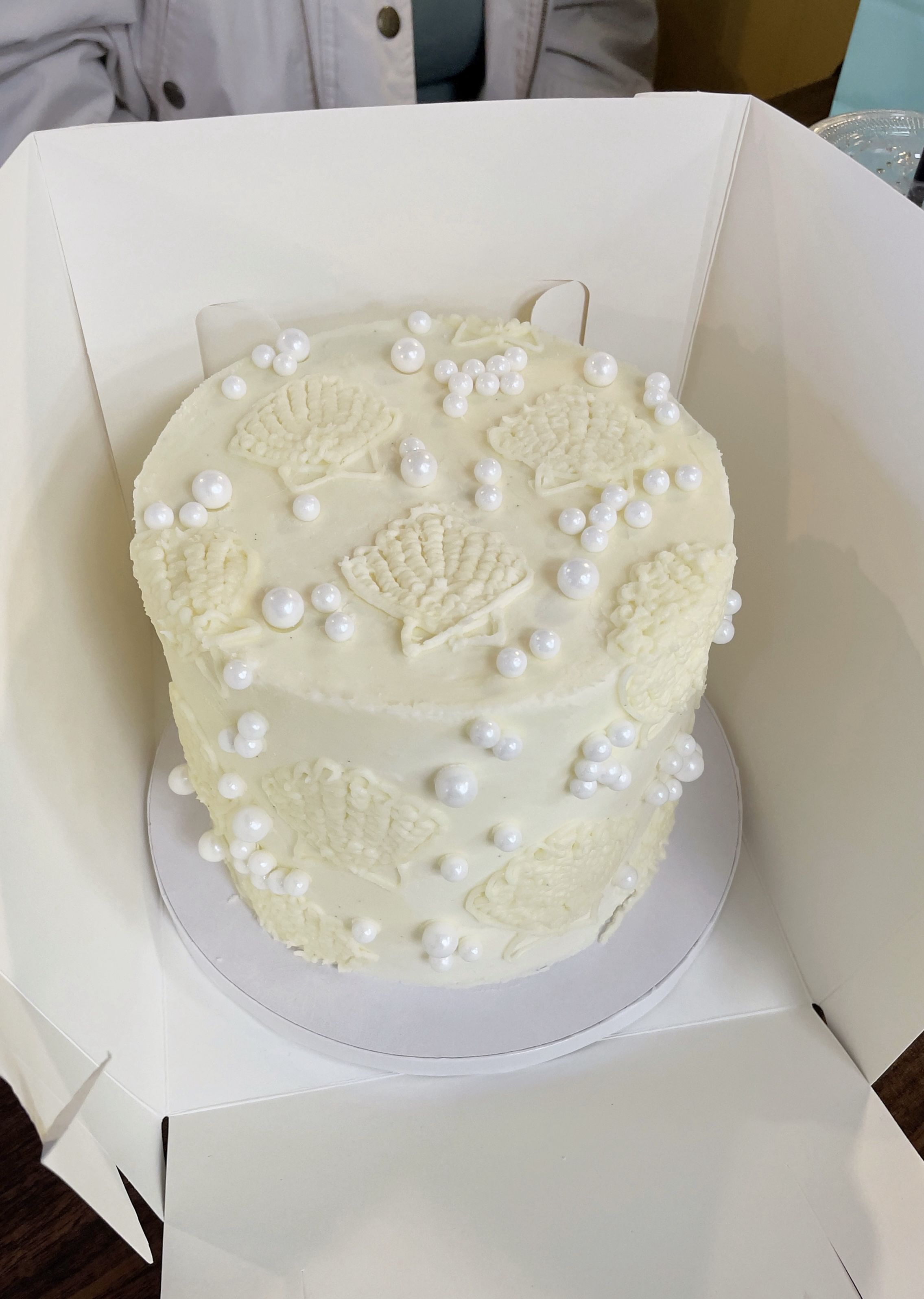 A white birthday cake decorated with shell-shaped designs and white pearl-like sprinkles, placed inside a white cake box.