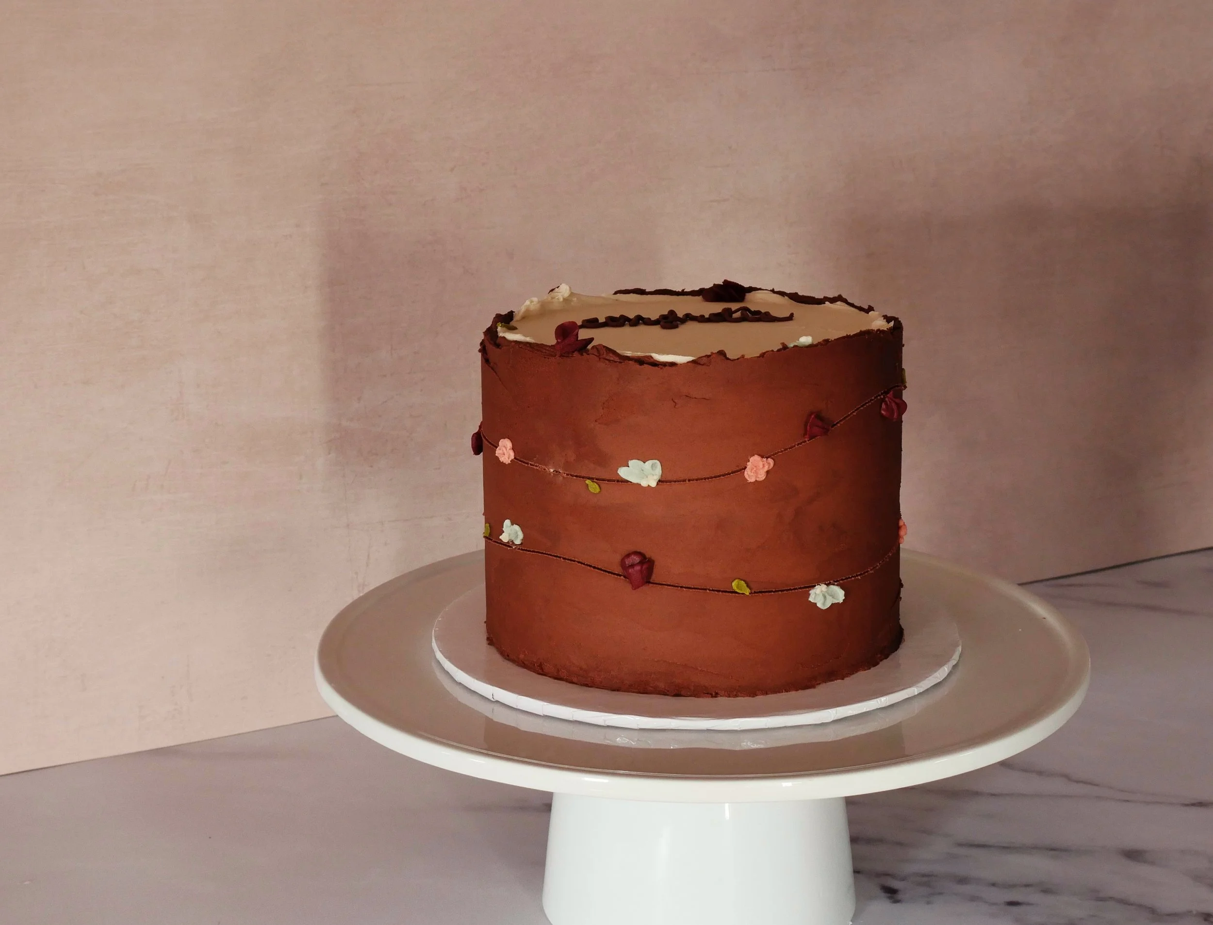 A decorated chocolate cake on a white pedestal cake stand, with pink, white, green, and red floral garland decoration and brown icing accents, placed on a marble surface against a beige background.
