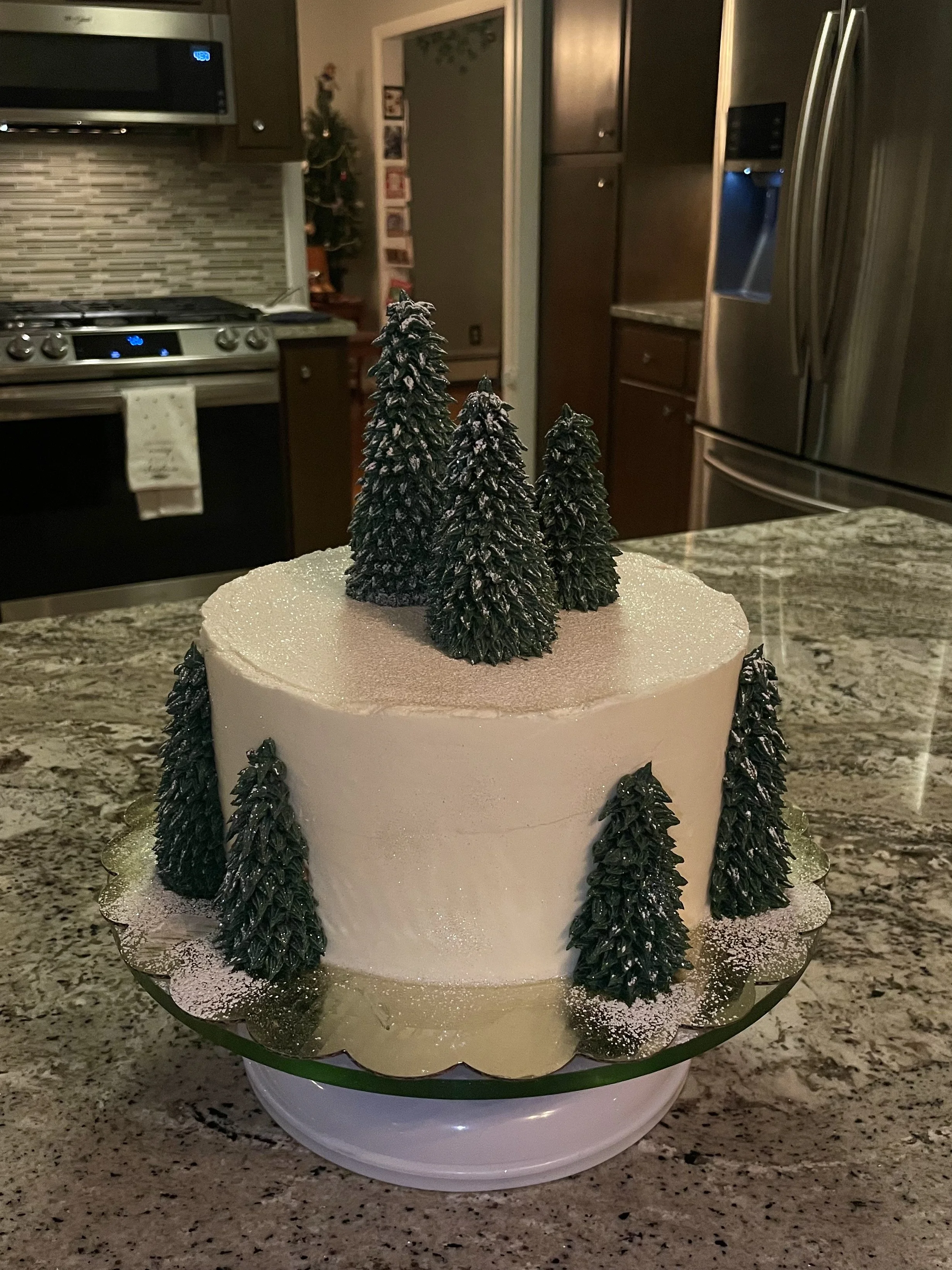 A white decorated cake with miniature evergreen trees on top and around the sides, placed on a gold cake board on a kitchen countertop.