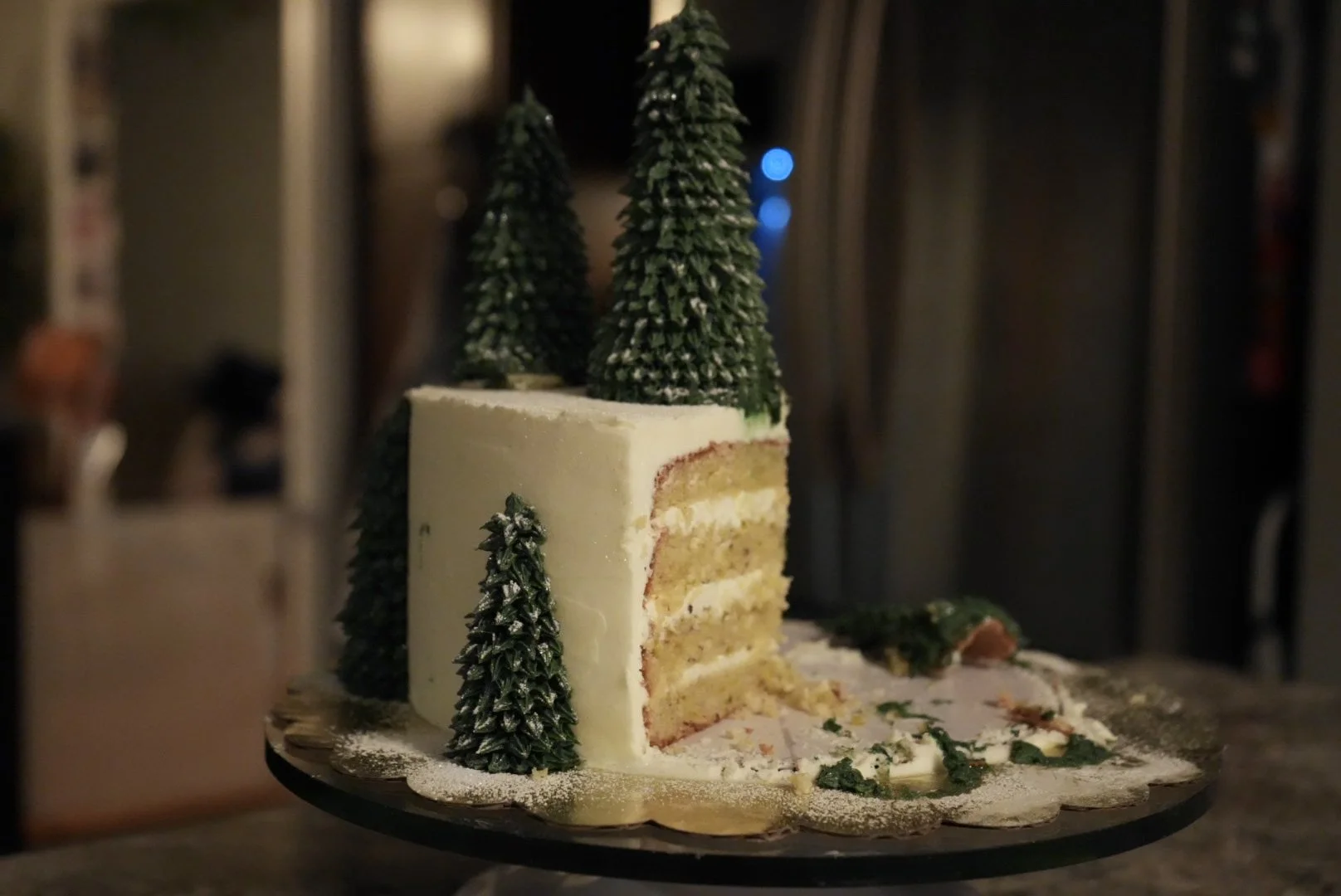 A partially cut Christmas-themed cake with green Christmas trees on top and around it, with pine and snow decorations.