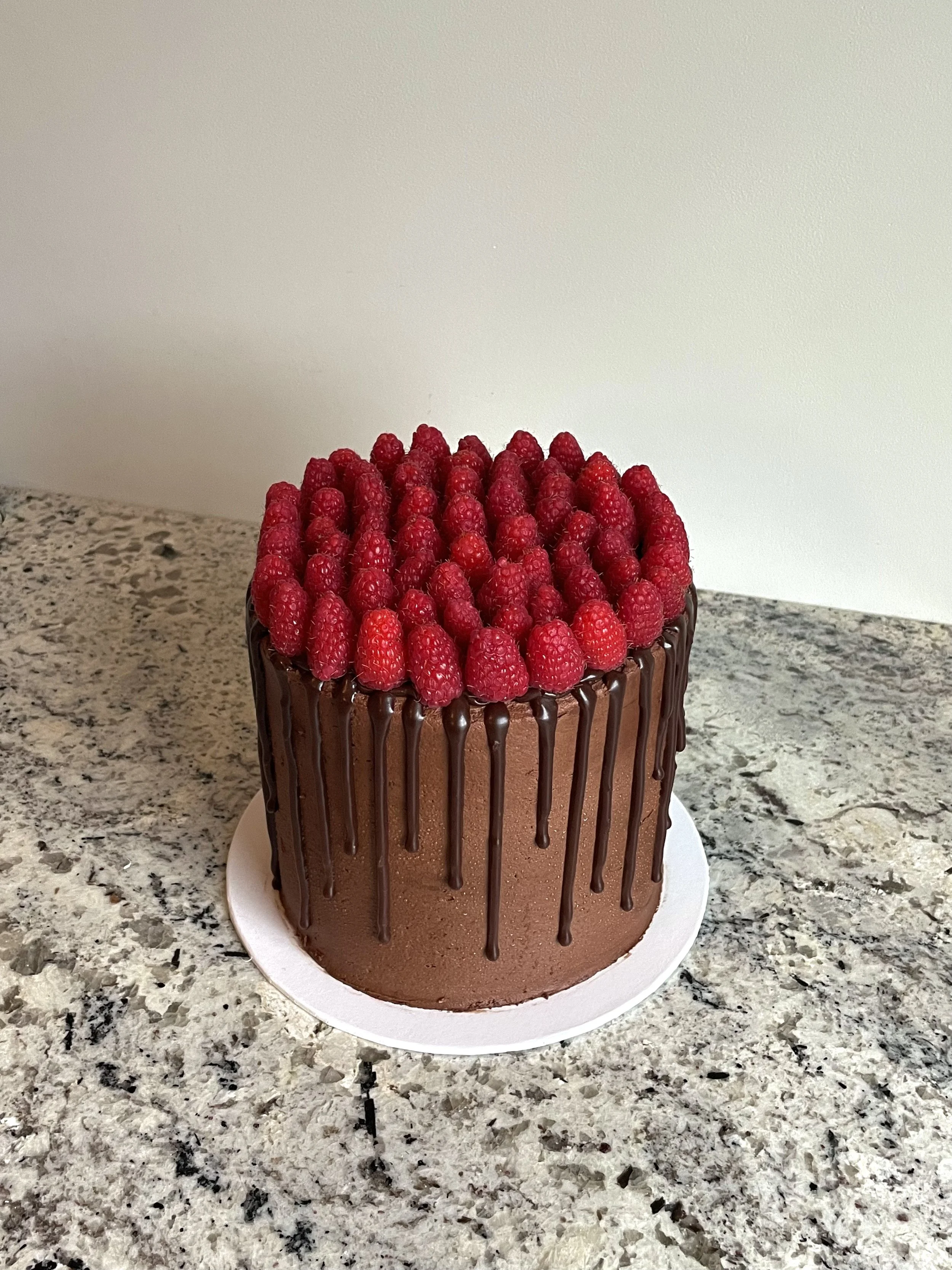 A chocolate cake decorated with a layer of raspberries and a chocolate drip glaze on a white cake board, placed on a speckled granite countertop.