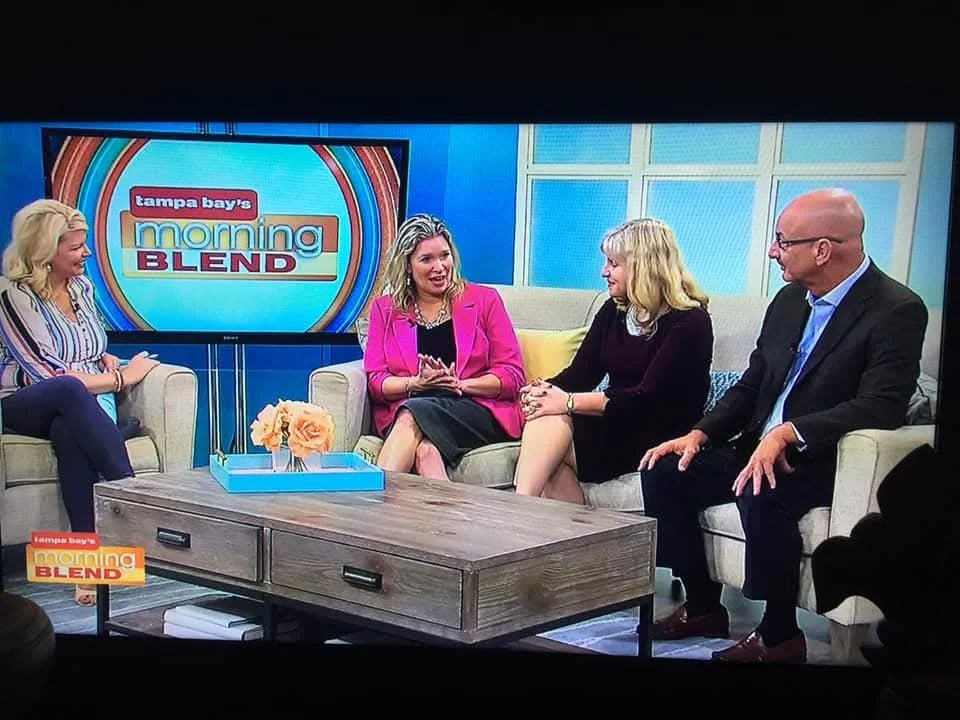 Four people sitting on sofas on a TV set with a large screen displaying the logo 'tampa bay's morning BLEND'. a woman on the left is wearing a striped shirt, and three women and one man are in the center and right, engaged in conversation.