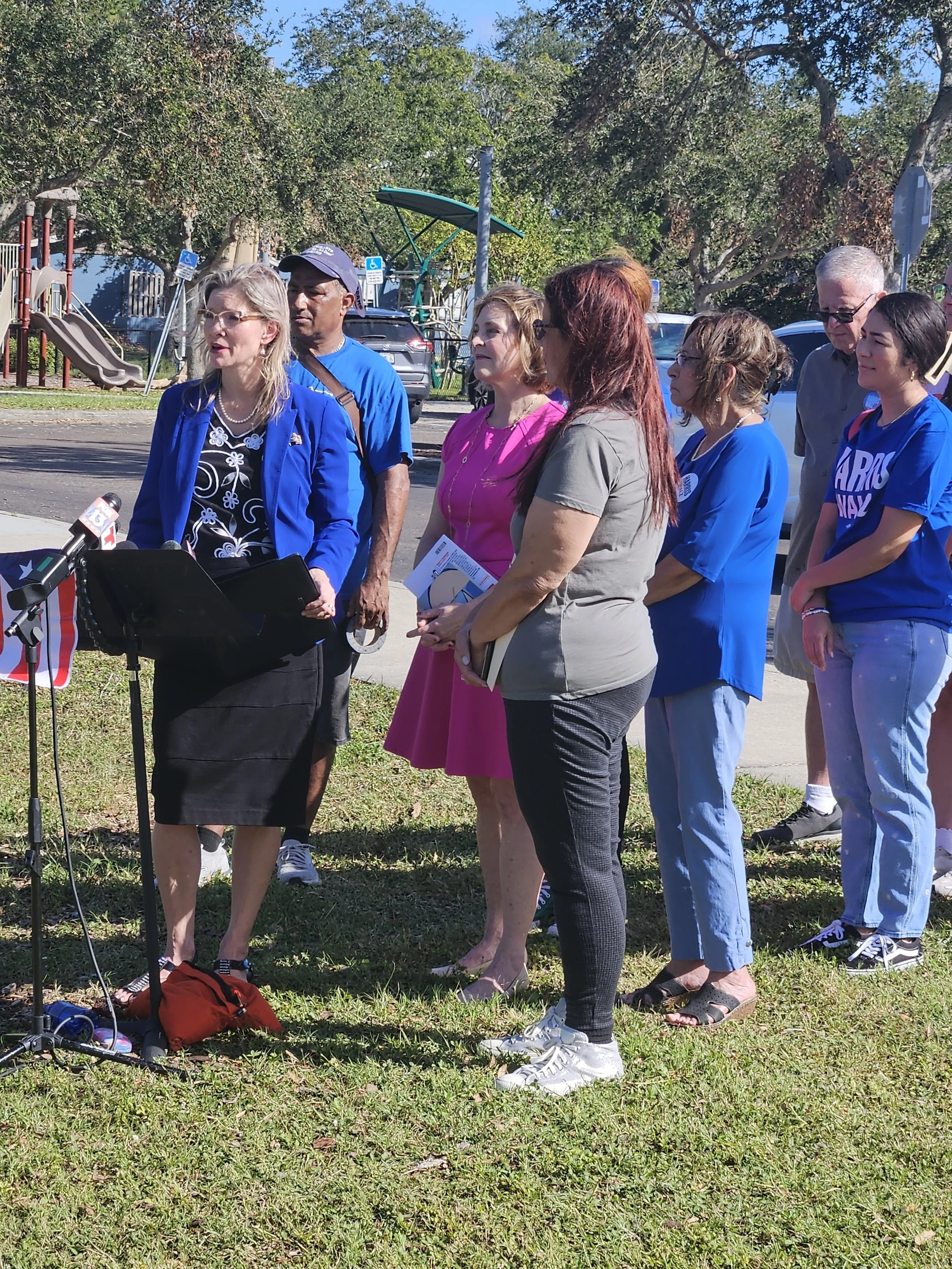 Group of people standing outdoors, possibly at a press conference or public event on a sunny day.