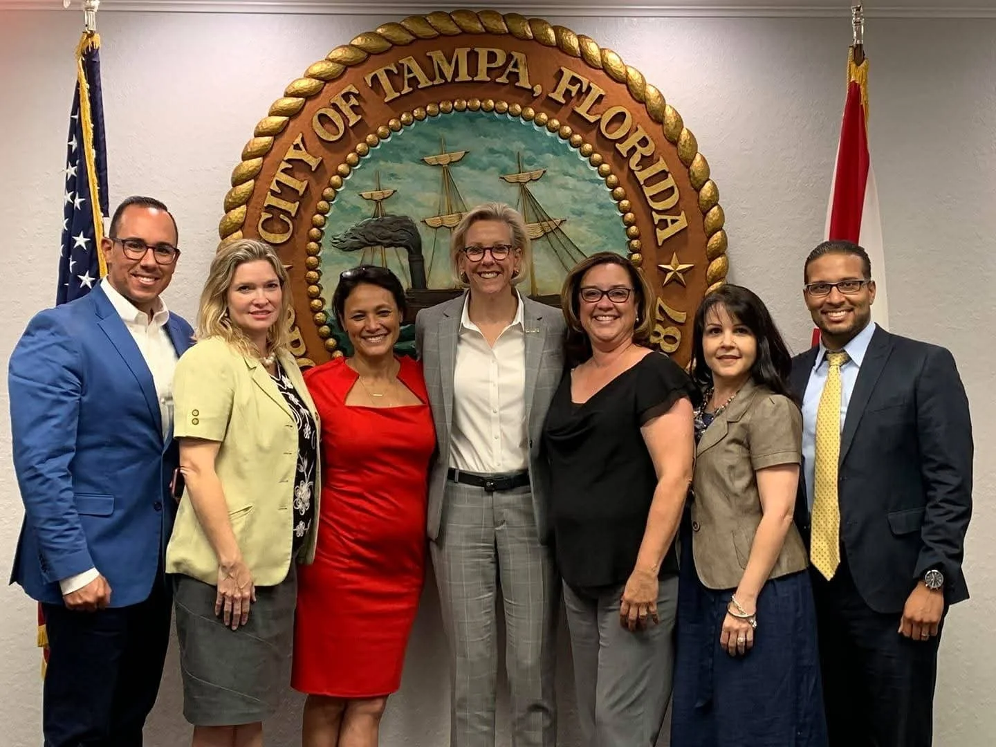 Group of seven people standing in front of the City of Tampa, Florida official seal, with flags on either side.