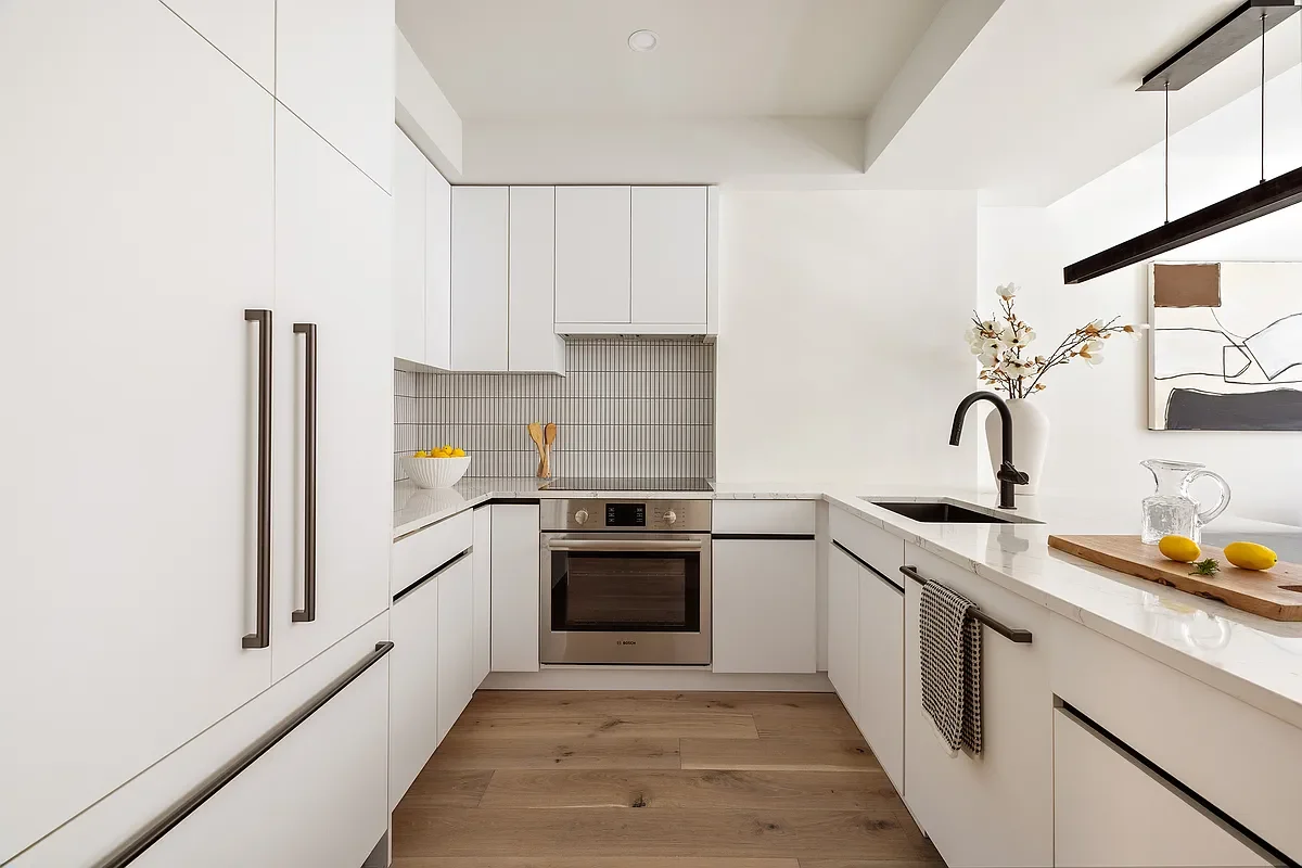 Modern white kitchen with gray accents and black hardware, featuring built-in appliances, a sink with a black faucet, and wooden accents on a countertop and cutting board.