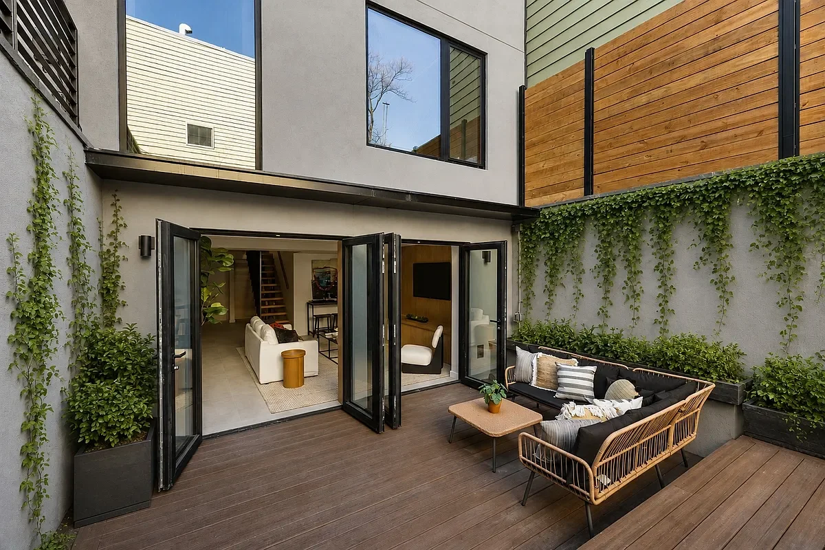 Modern outdoor patio with black metal and wooden furniture, plants, and open glass doors leading inside a house.