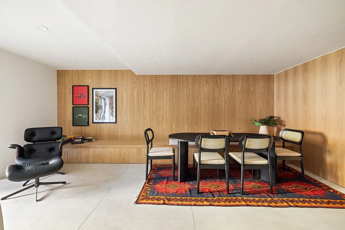 Modern living room with a black lounge chair, a round black dining table with six chairs, a wooden wall panel, framed artwork, a red patterned rug, and a potted plant.