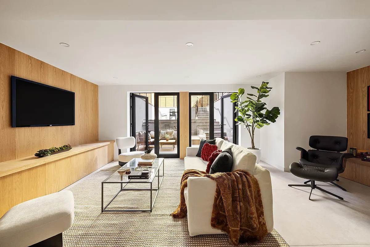 Modern living room with white sofa, black accent pillows, and a rust-colored throw blanket, glass coffee table, built-in wooden shelf, large window doors, potted plant, black lounge chair, and wall-mounted flat-screen TV.