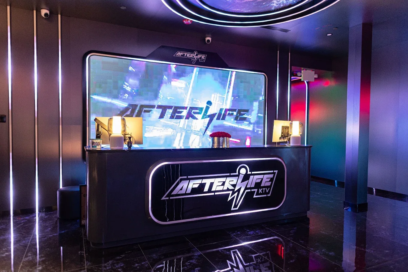 Reception desk with illuminated sign for Afterlife KTV, featuring microphones and desktop lamps, with a large screen behind displaying the logo and a colorful, vibrant background.