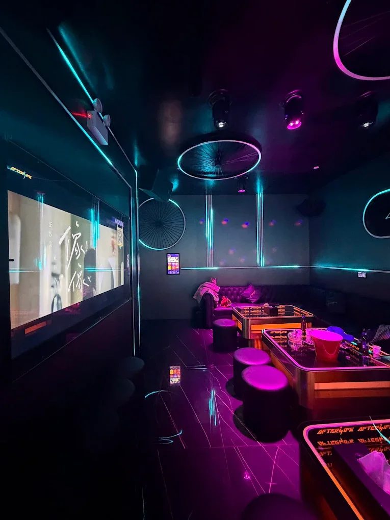 A dimly lit room with neon lights, a large flat-screen TV, a sofa, and tables with stools, decorated with glowing ping pong paddles and bicycle wheel wall art, creating a modern, lounge-like atmosphere.