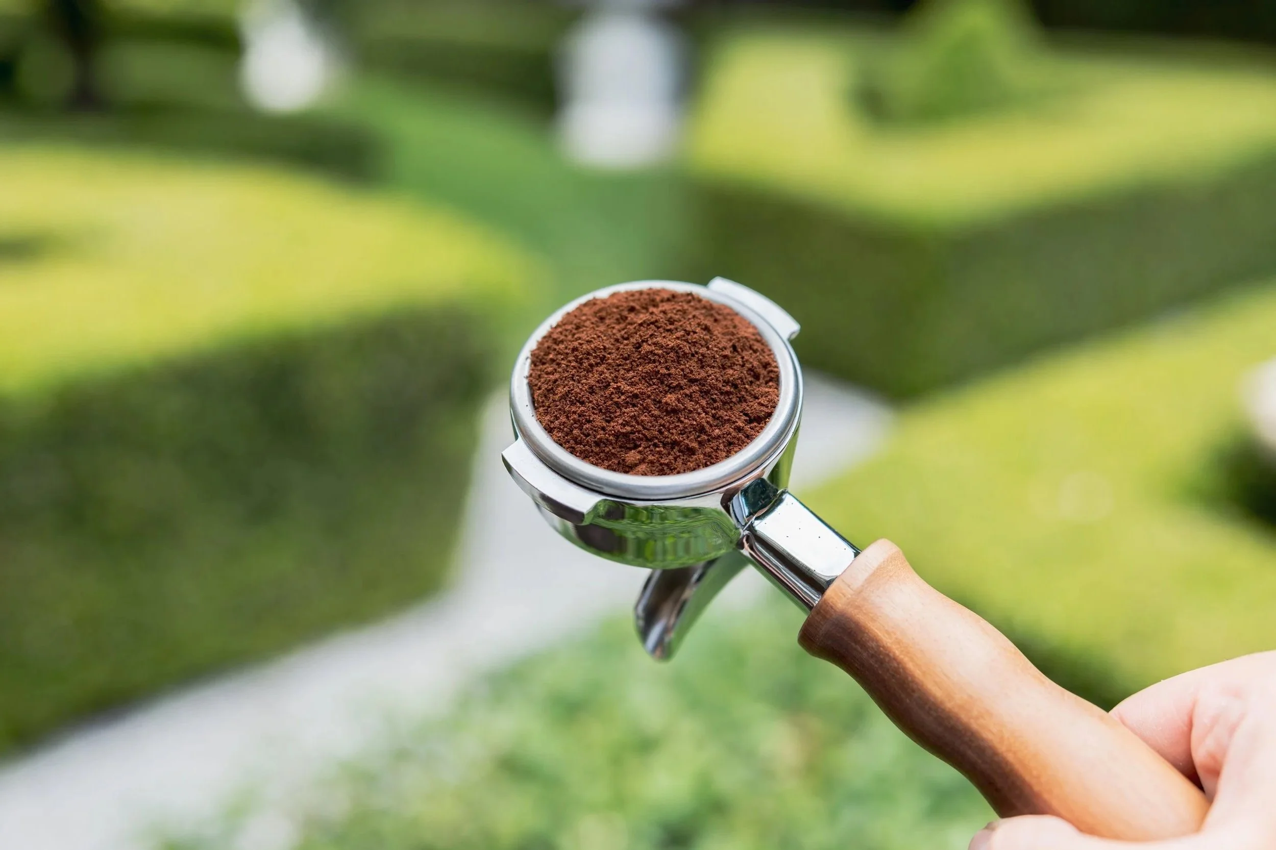 Close-up of a portafilter with ground coffee, held by a hand outdoors with green trees and grass in the background.