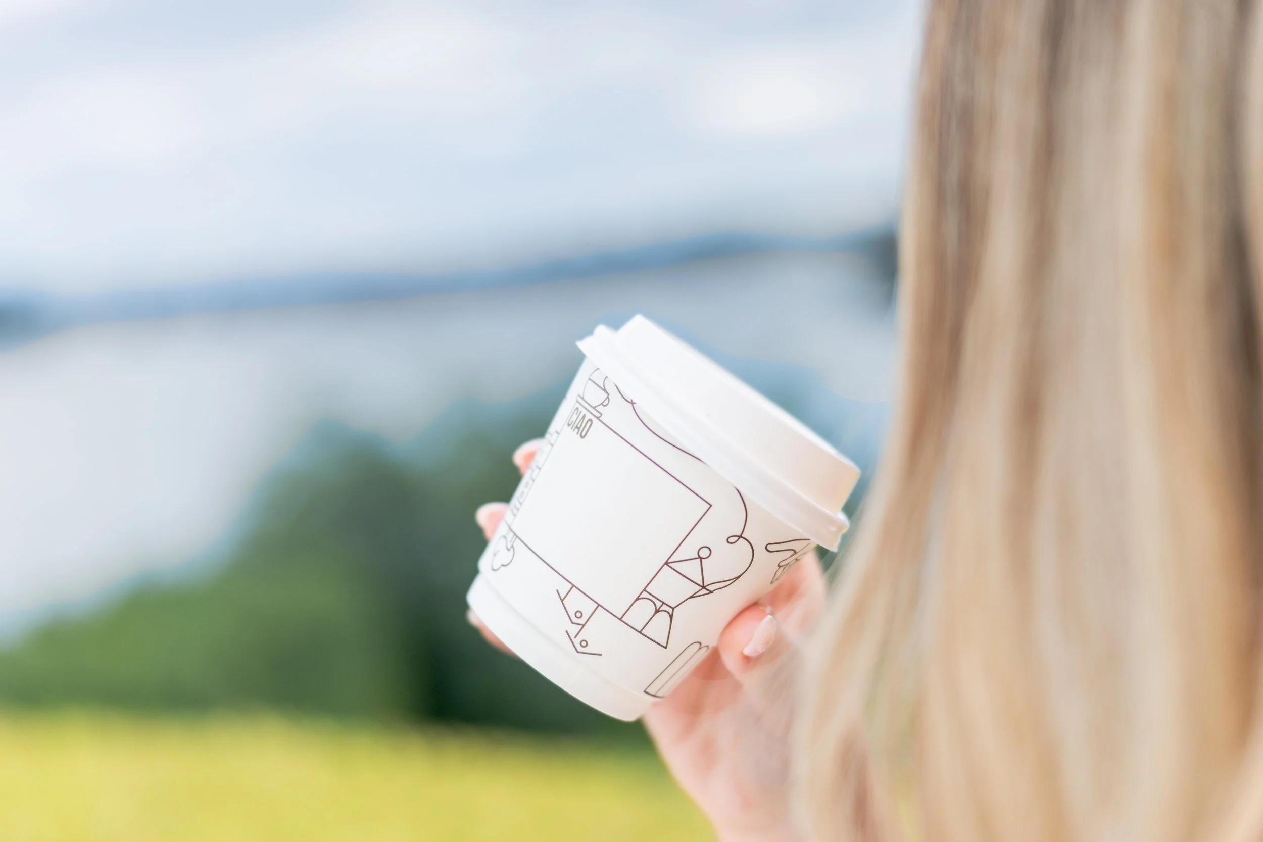 Person holding a white disposable coffee cup outdoors with a blurred green and blue background.