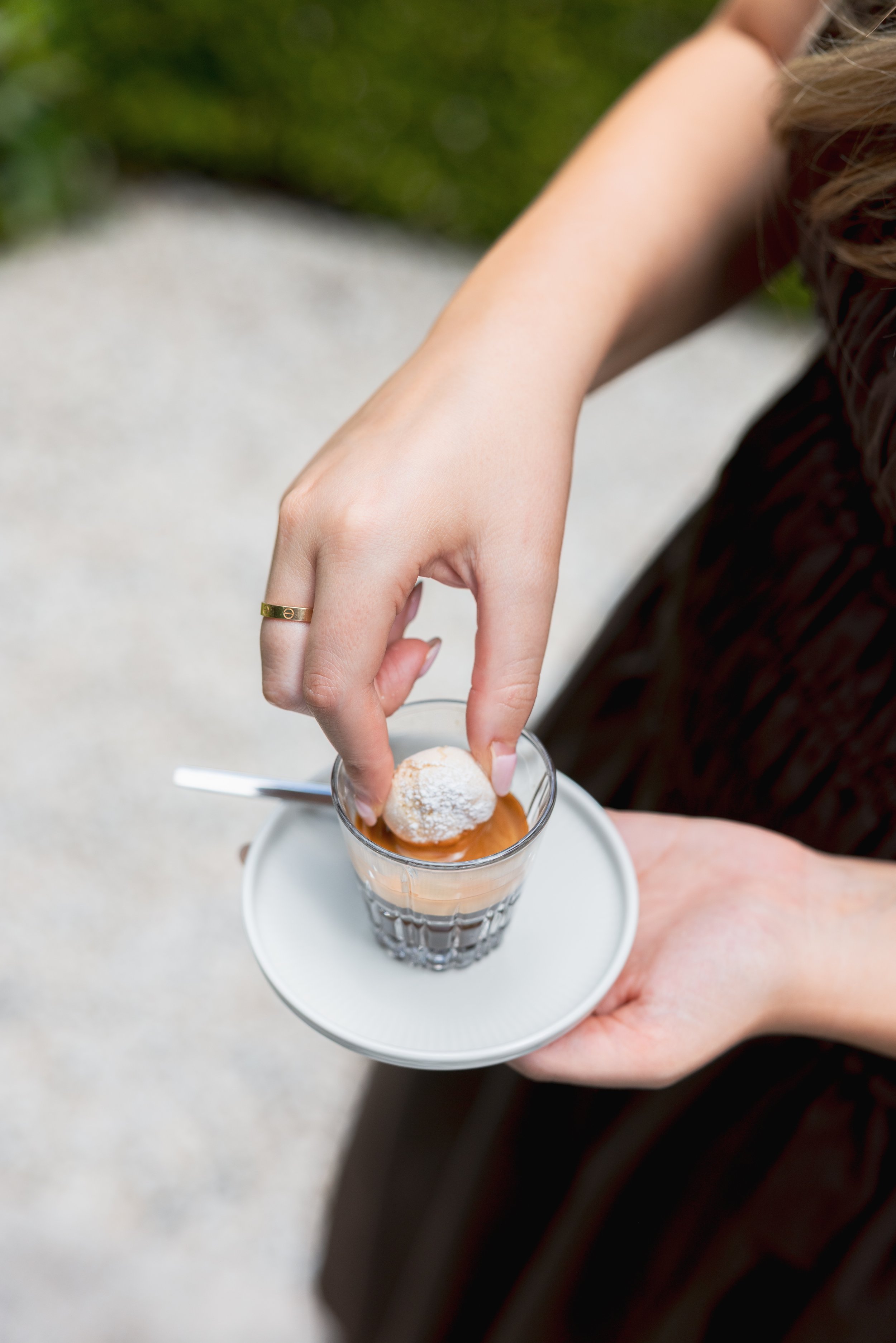 A person holding a glass of coffee or dessert with a small powdered sugar-covered pastry on top, on a white saucer.