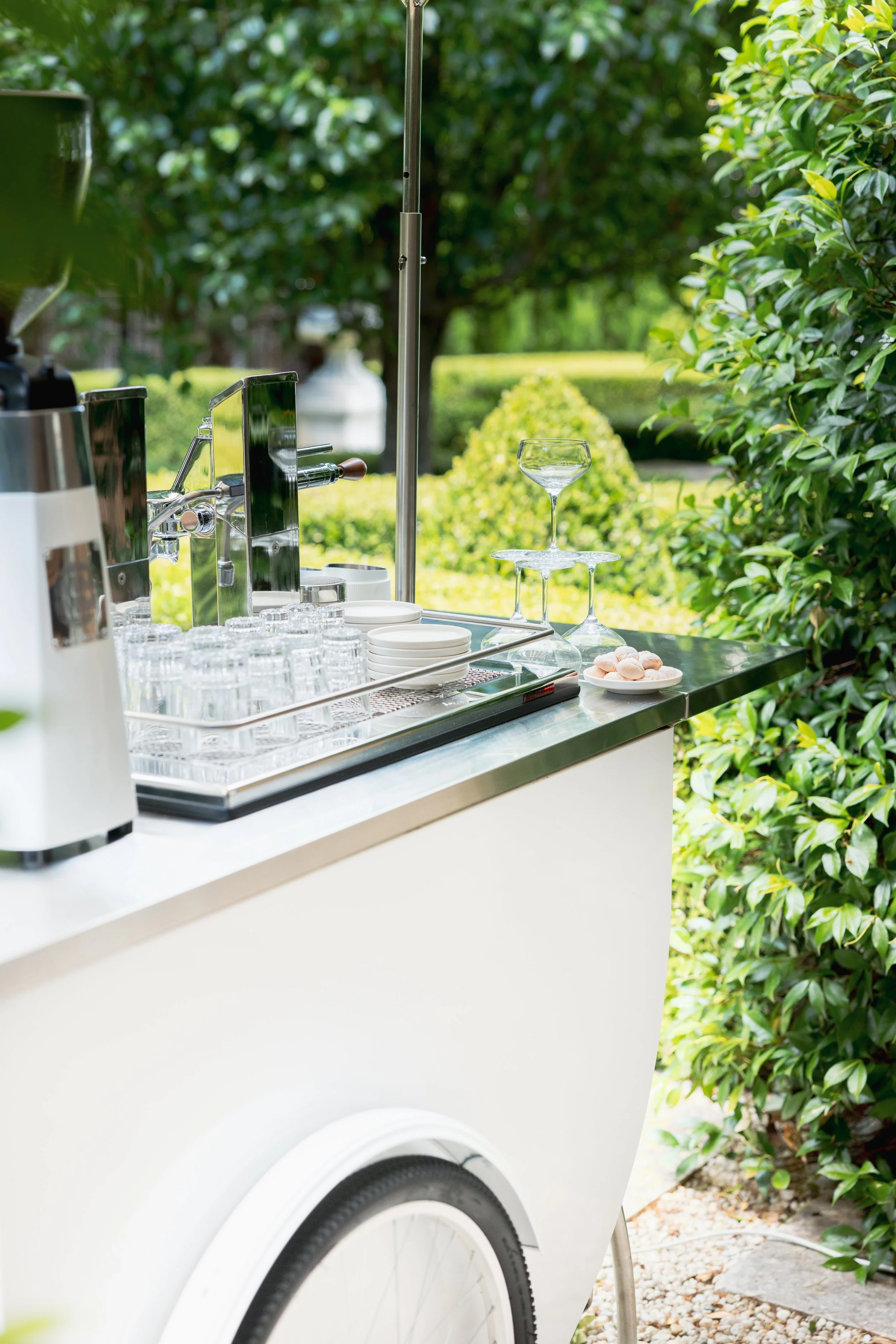 A mobile beverage bar with glasses, plates, and a drink dispenser, set outdoors with green bushes and trees in the background.